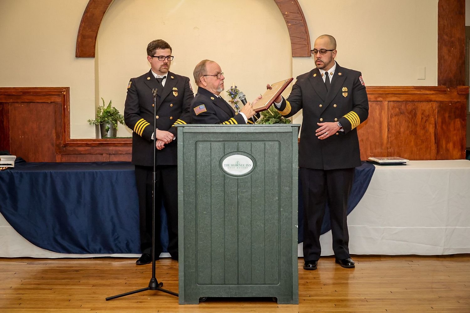 Three military officers in uniform stand at a podium during a formal ceremony in an elegant hall.