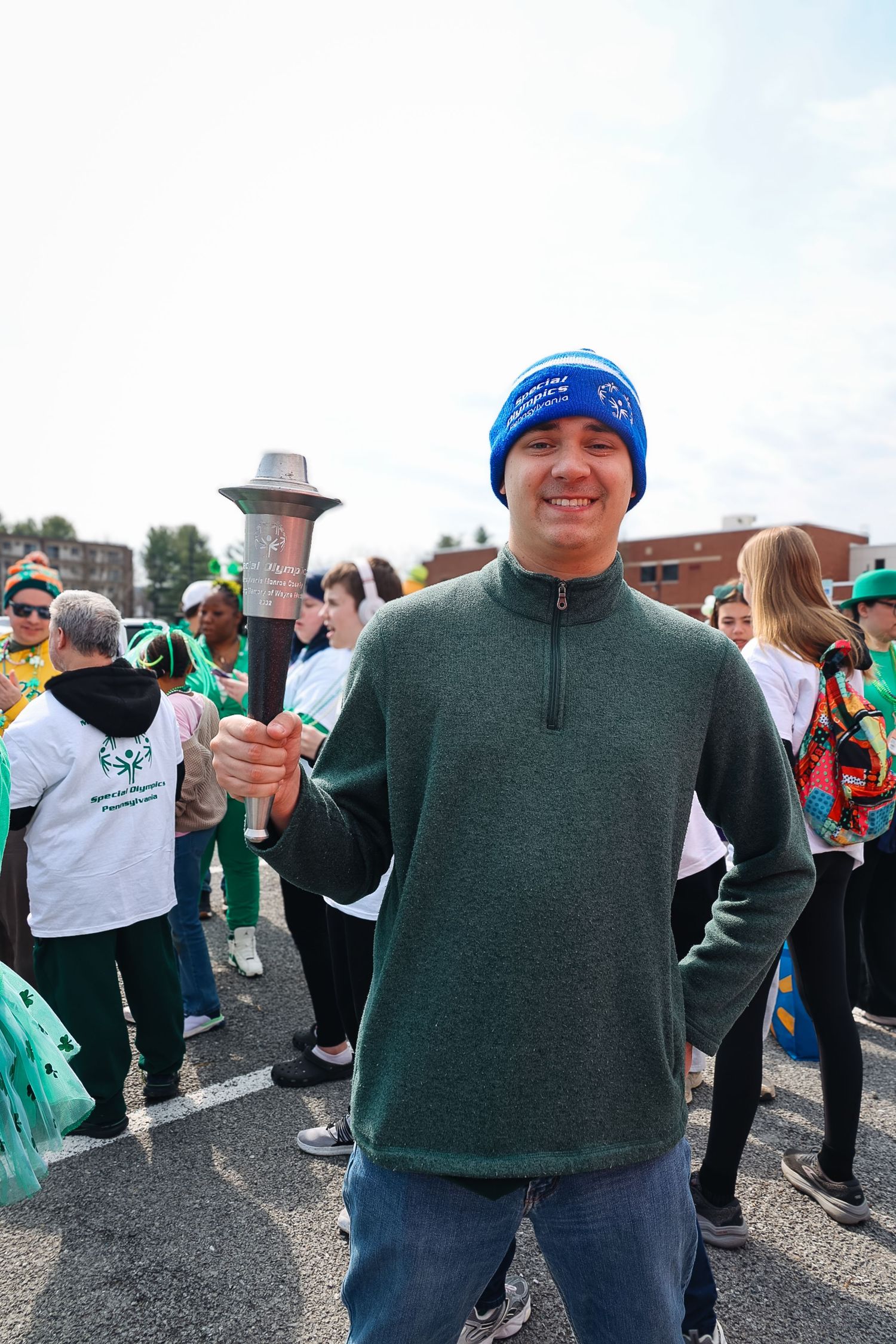 Man in blue beanie and green pullover holds a torch at an outdoor charity walk event.