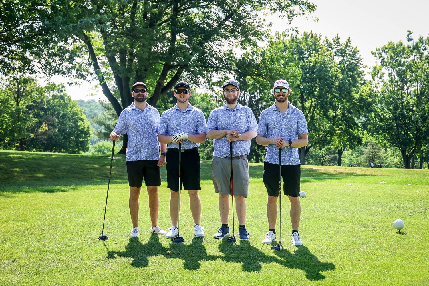 Four golfers posing together on a sunny golf course, holding clubs and smiling in matching blue polo shirts.