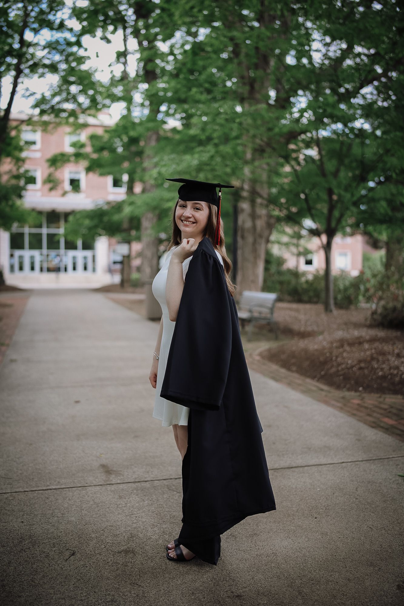 A smiling woman in a black graduation cap and gown poses on a tree-lined campus walkway.