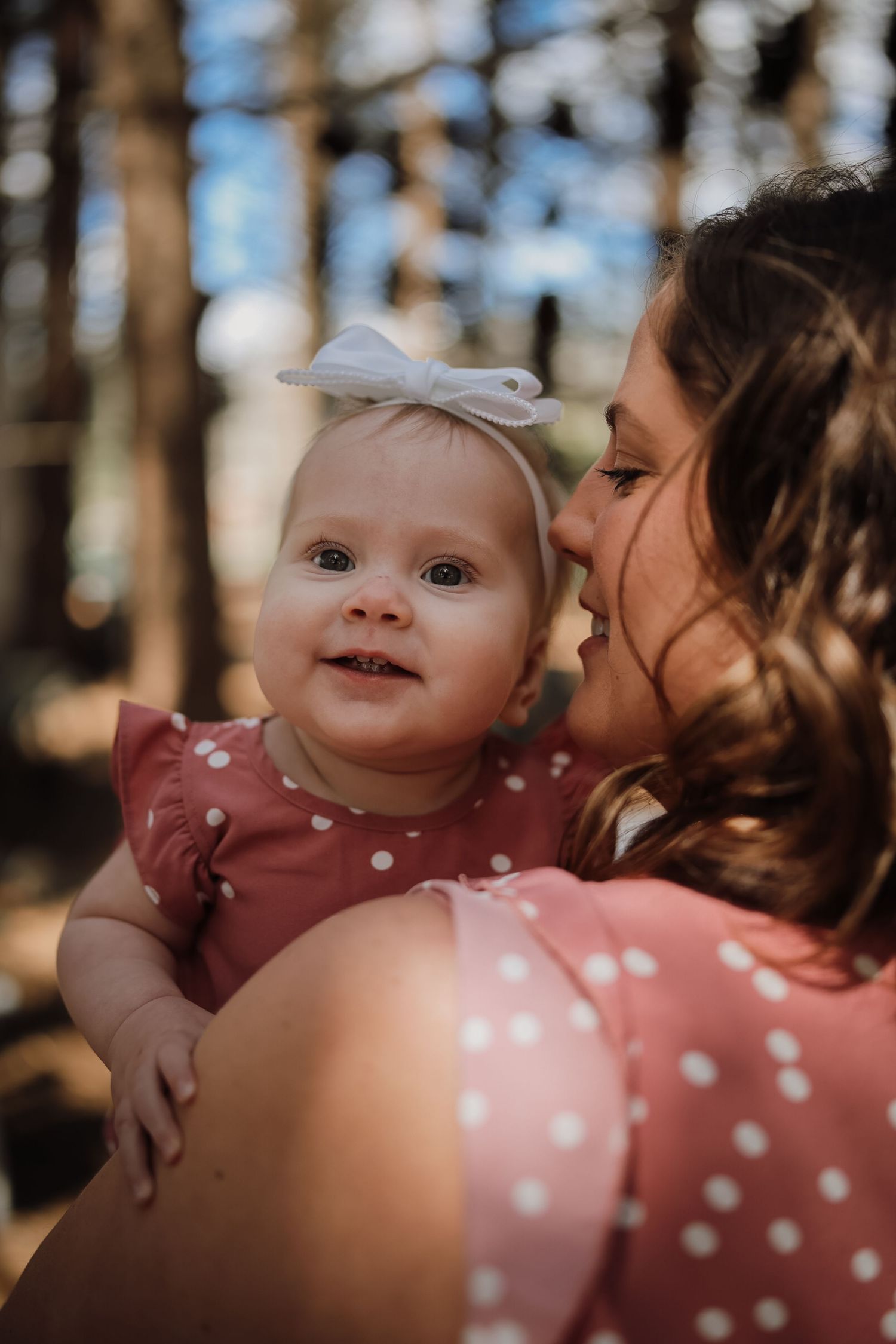 A smiling baby in a red polka dot dress with a white bow headband being held by a woman in a matching dress outdoors.