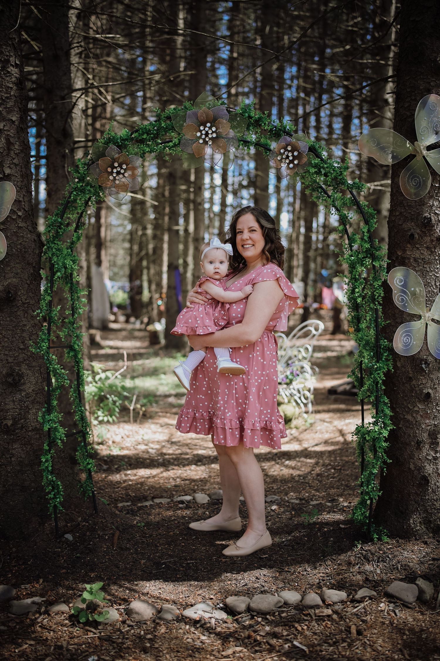 Mother in pink dress holds baby in matching outfit under a green floral arch in a woodland setting.