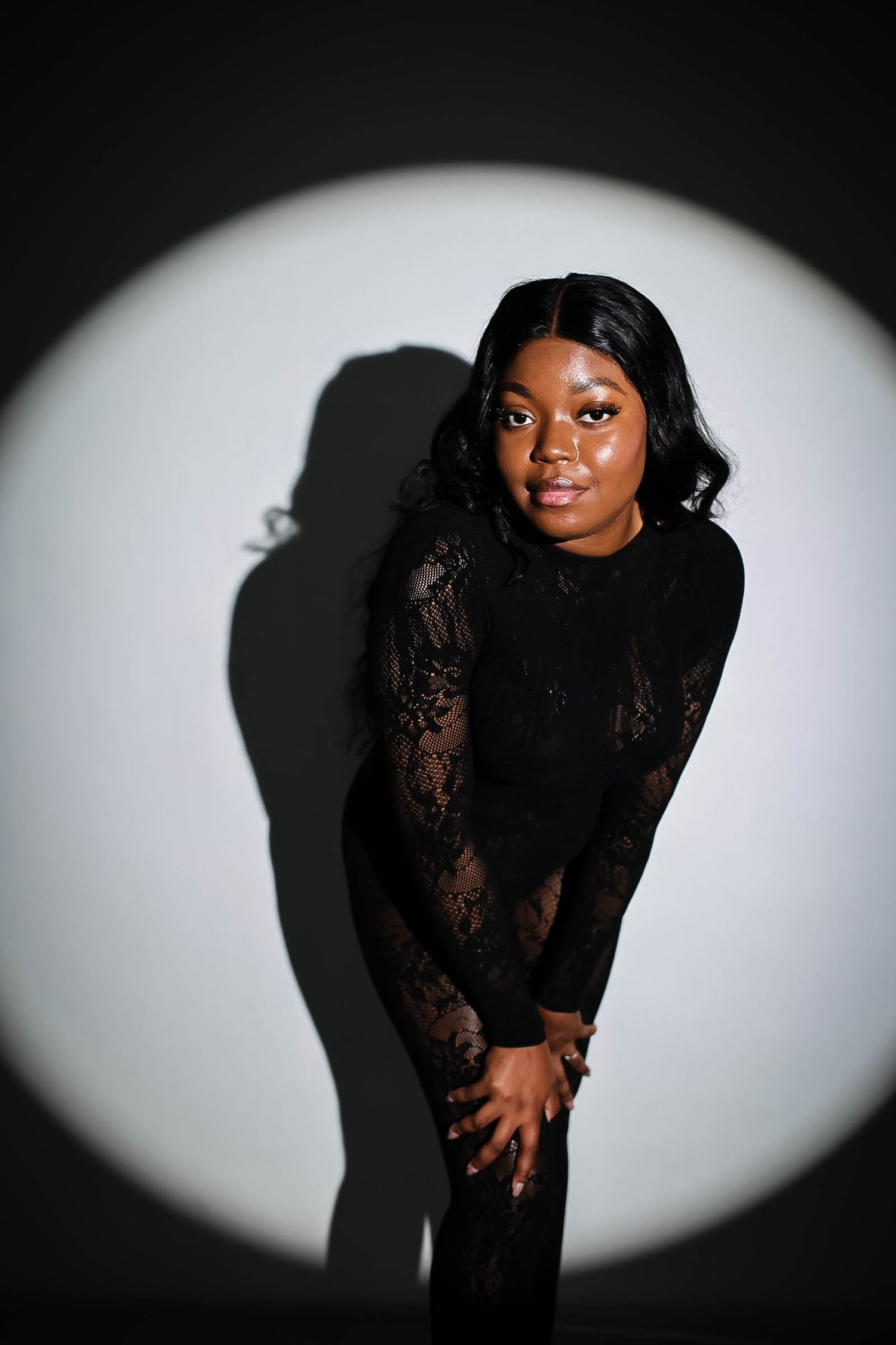 Young woman in black lace outfit poses confidently under dramatic spotlight against gray background.