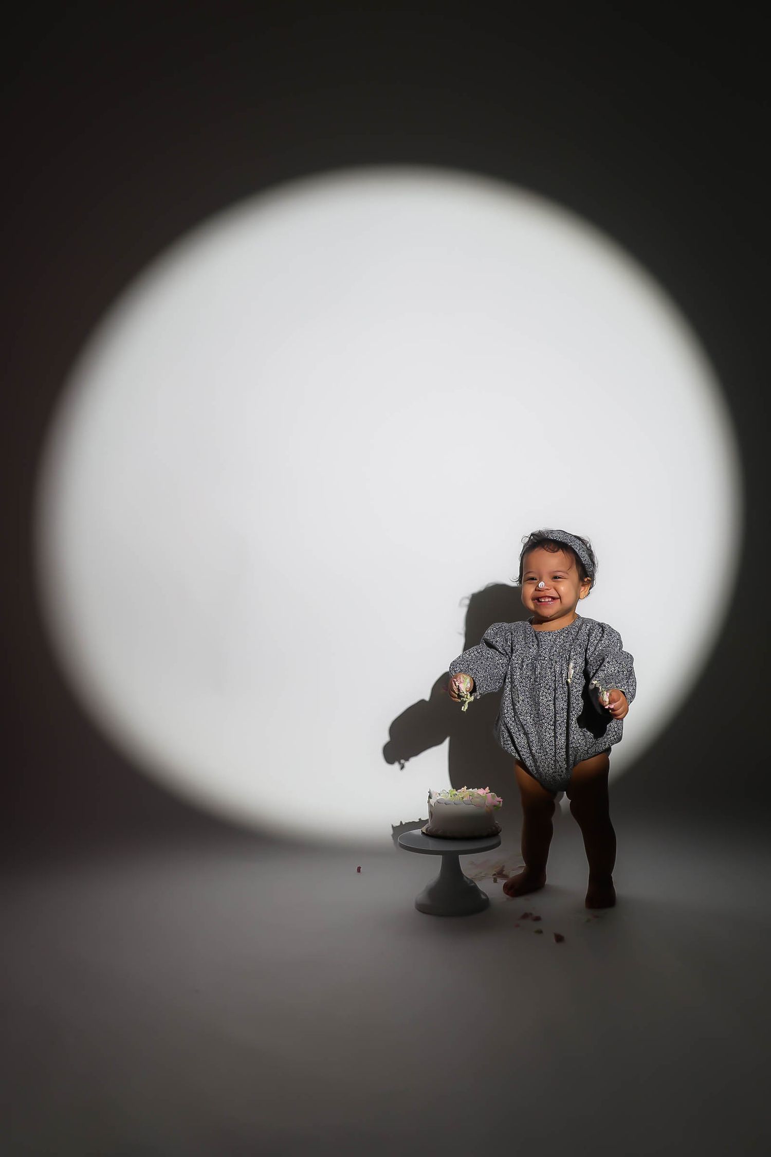 Happy toddler in gray romper laughing beside a smashed cake under a circular spotlight in a studio.