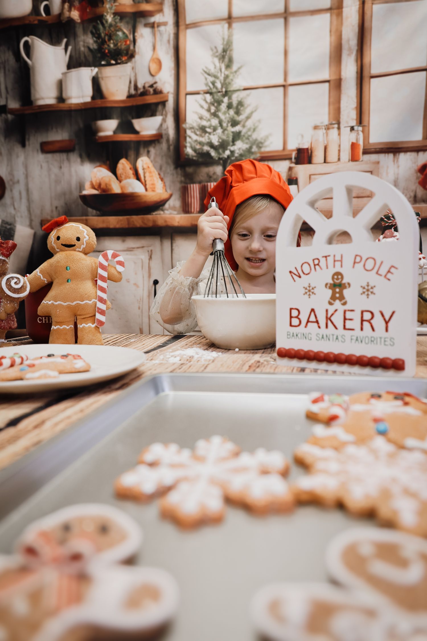 Child in chef hat baking Christmas cookies at North Pole Bakery with gingerbread decorations.