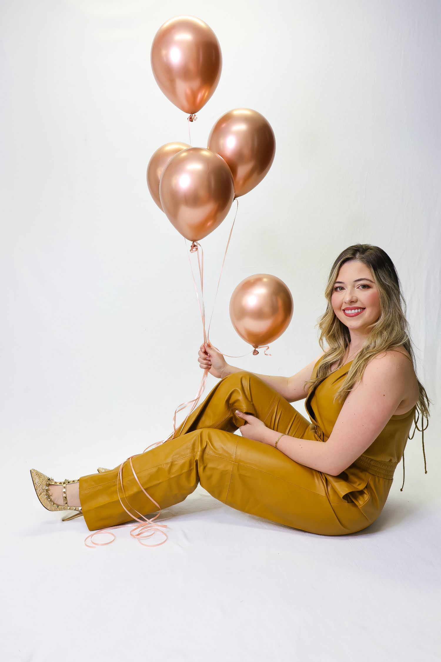 Woman in mustard yellow jumpsuit sitting on white floor holding rose gold metallic balloons, smiling at camera.
