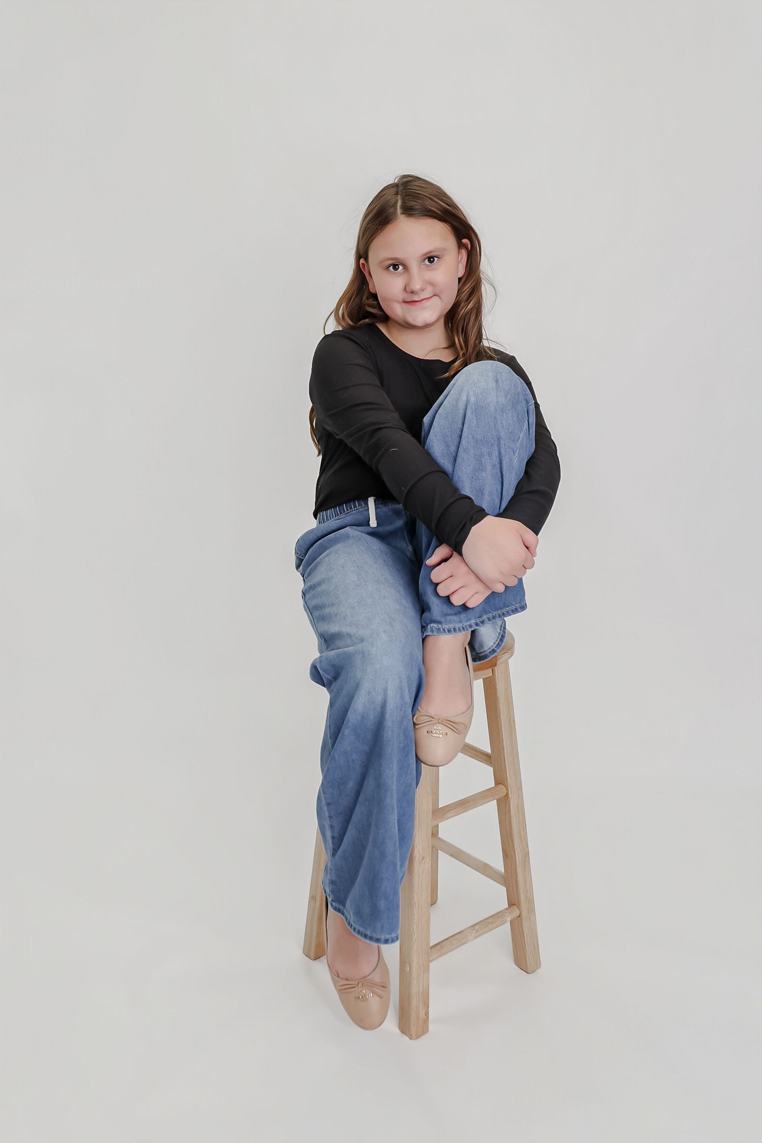 A young girl sits on a wooden stool, wearing a black top and blue jeans, smiling at the camera against a white background.