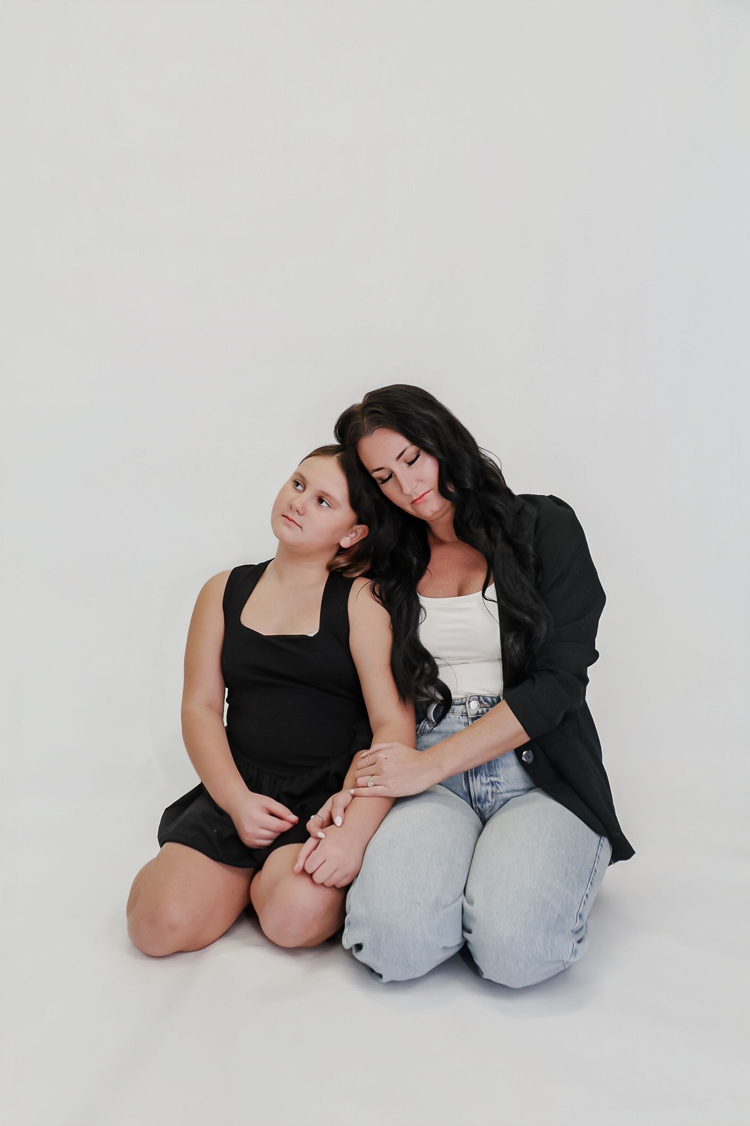 A woman with dark hair in a black blazer sits on the floor embracing a young girl in a black dress against a white background.