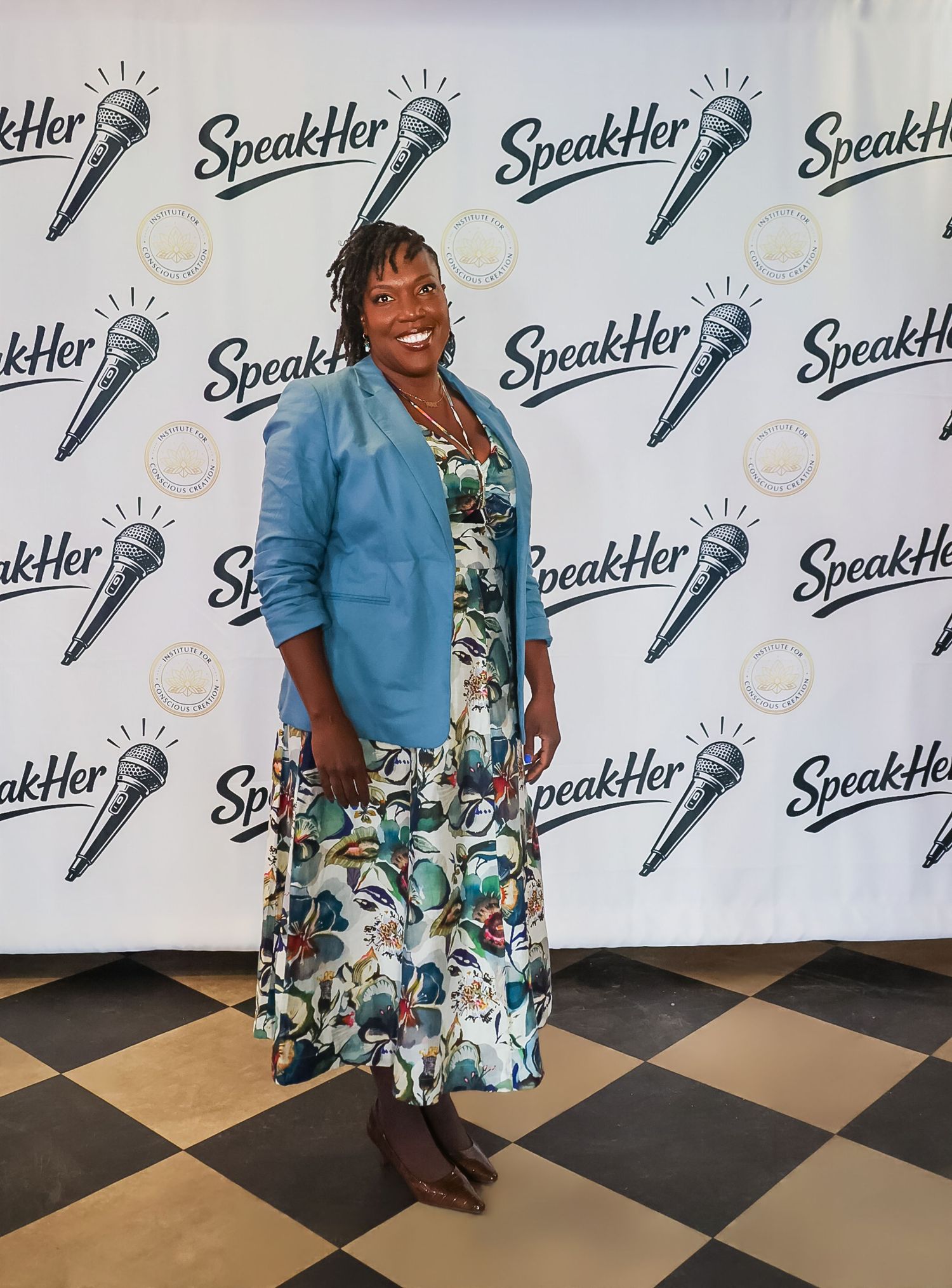 Woman in floral dress and blue blazer poses at SpeakHer event backdrop with microphone logos.