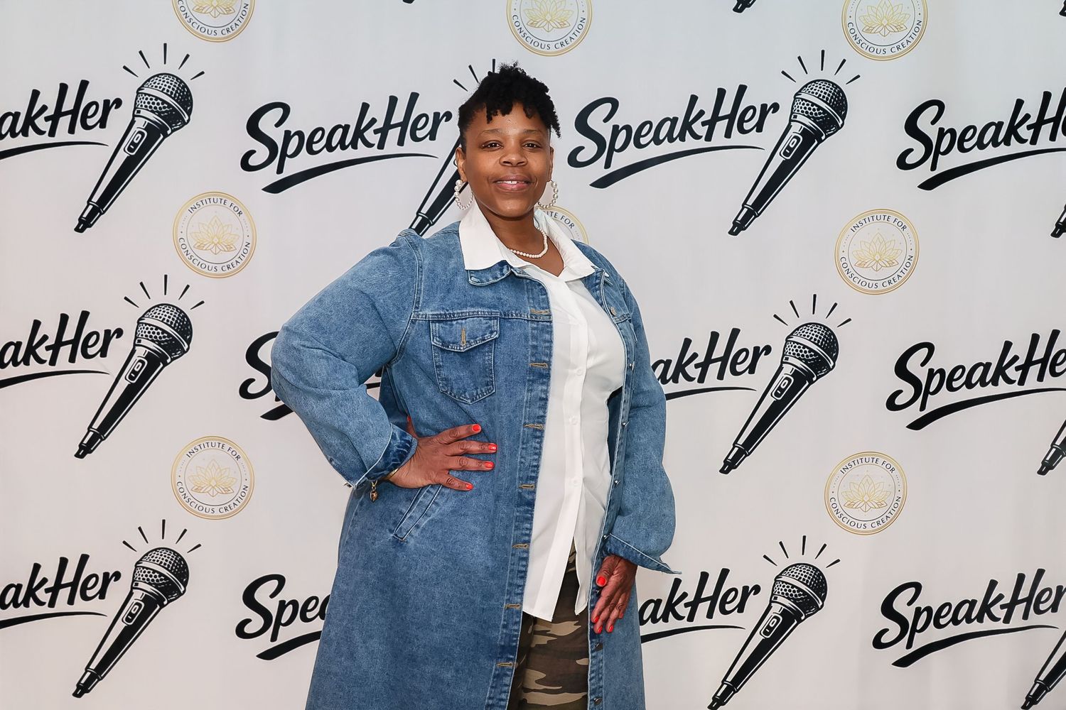 Woman in denim jacket poses at SpeakHer event backdrop with microphone logo.