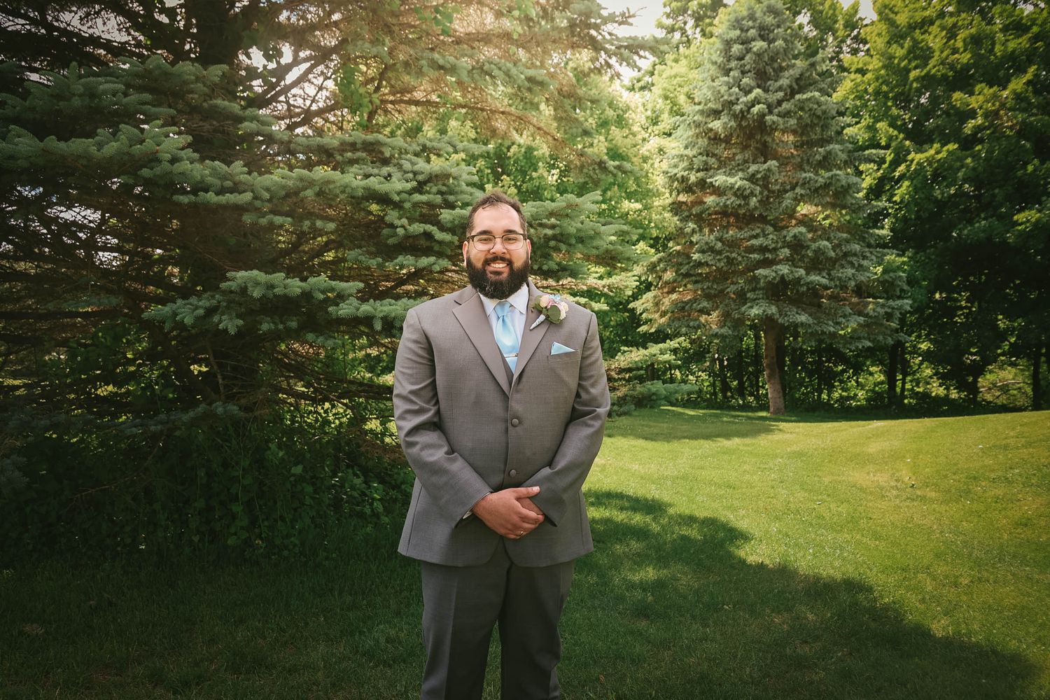 A bearded groom in a gray suit with light blue tie stands on green lawn surrounded by lush trees at an outdoor wedding.