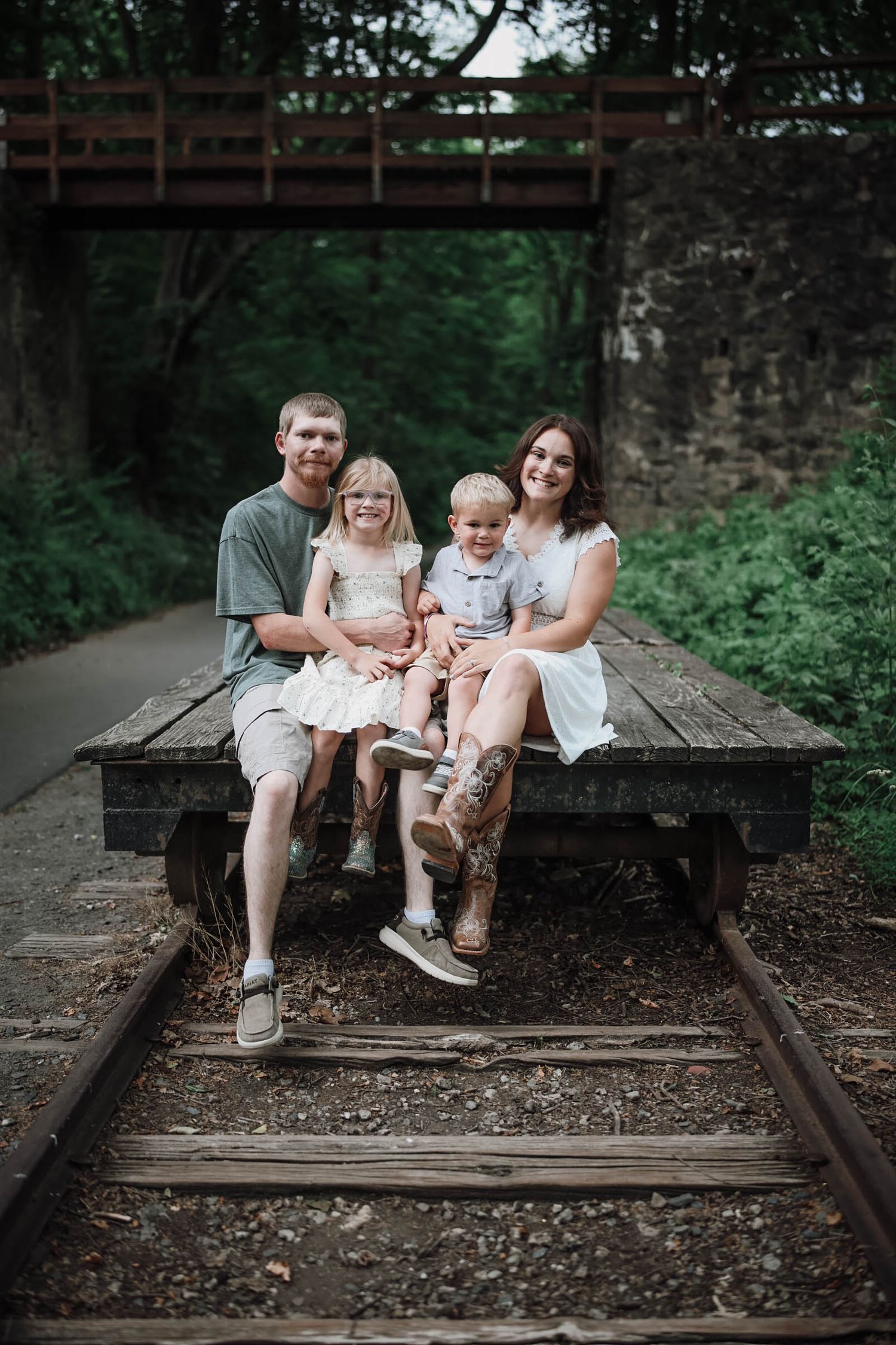 Family of four smiling on vintage rail cart under bridge surrounded by lush greenery.