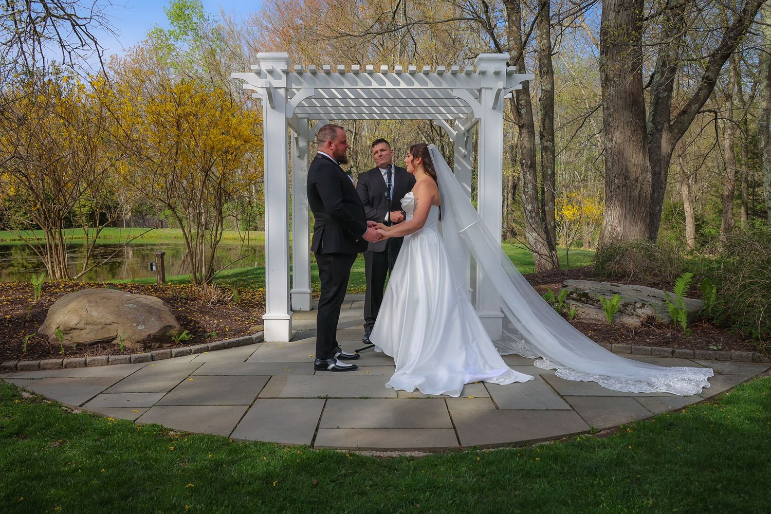 Bride and groom exchange vows under white pergola surrounded by lush greenery in outdoor spring wedding ceremony.