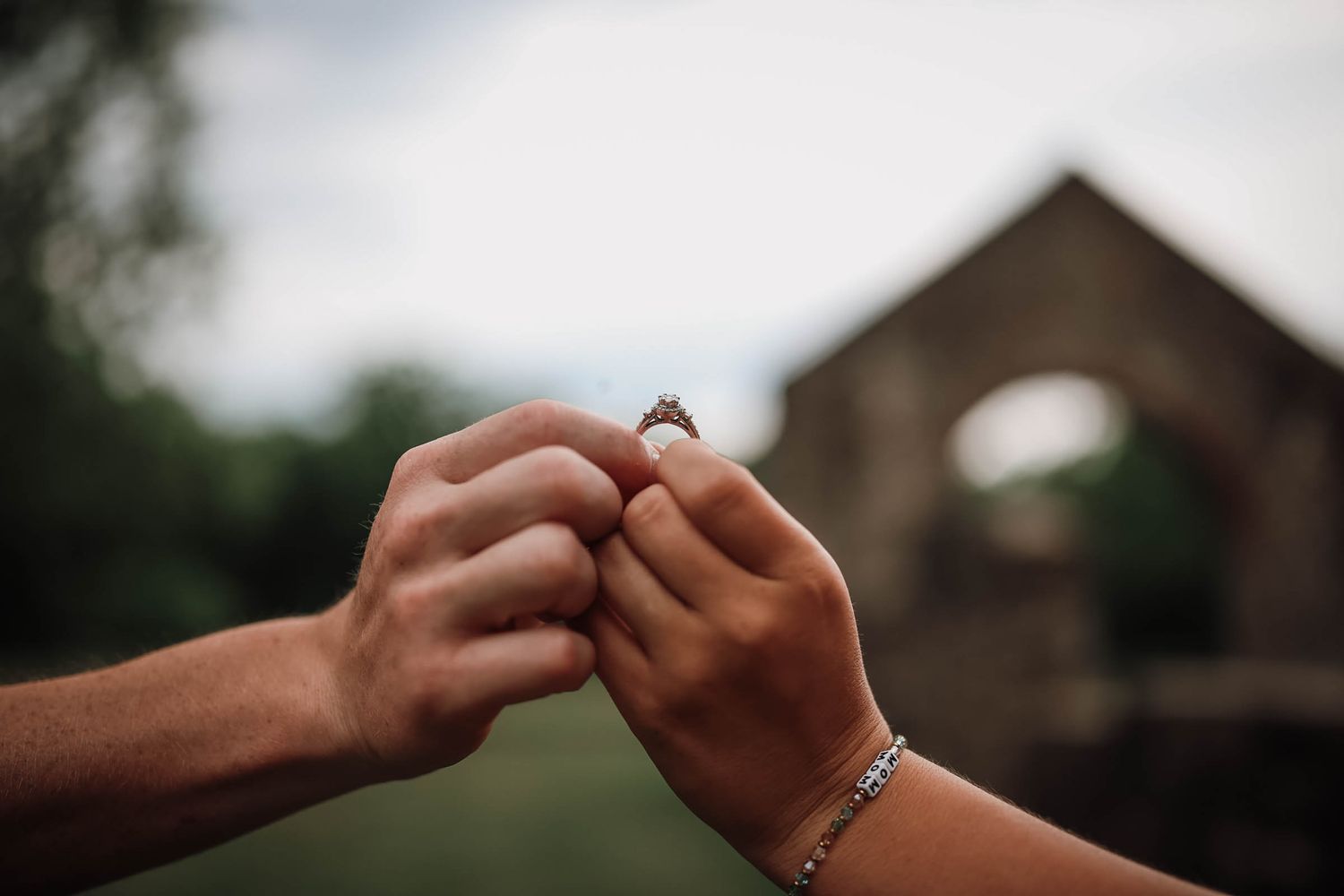 Two hands holding a diamond engagement ring together outdoors near a rustic stone building.