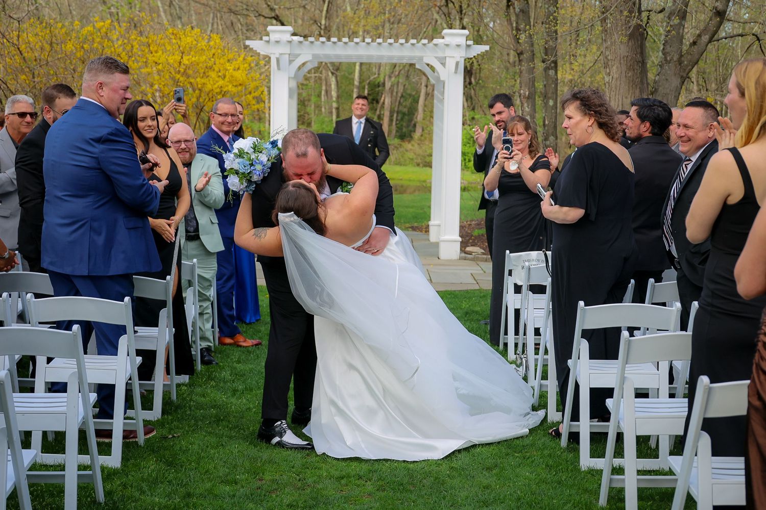 Groom dips bride for a kiss at outdoor wedding ceremony with white chairs and floral arbor backdrop.
