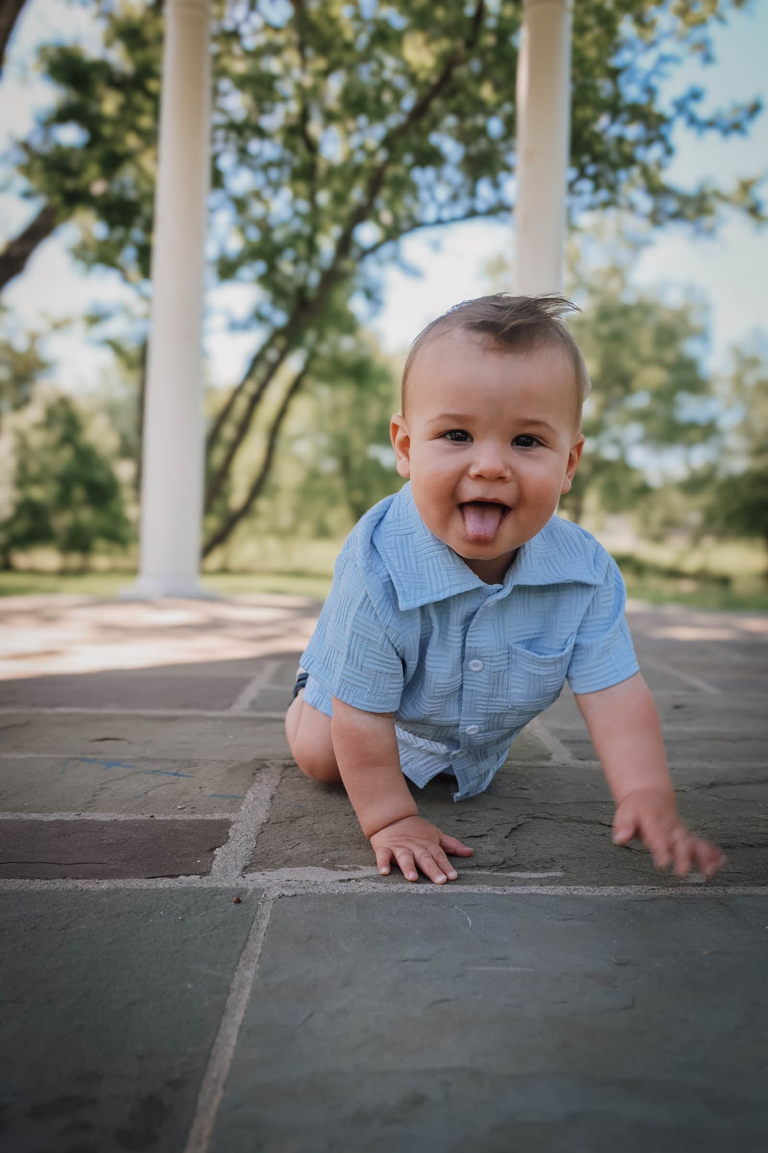 A joyful baby boy in a blue romper sticks out his tongue while crawling on a brick patio outdoors.