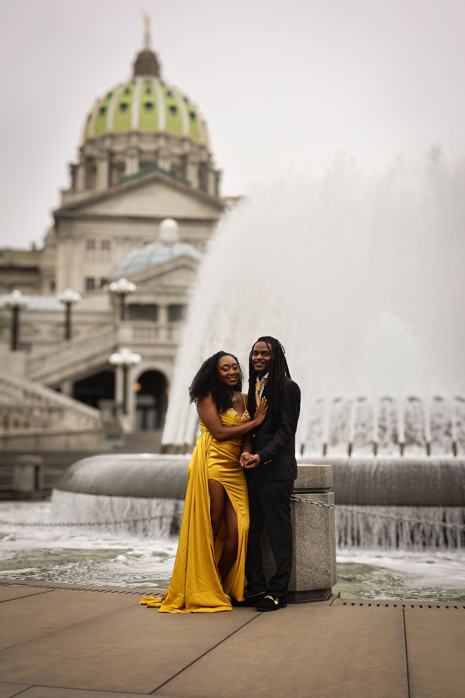A couple in formal attire poses together in front of the Pennsylvania State Capitol building and fountain in Harrisburg.