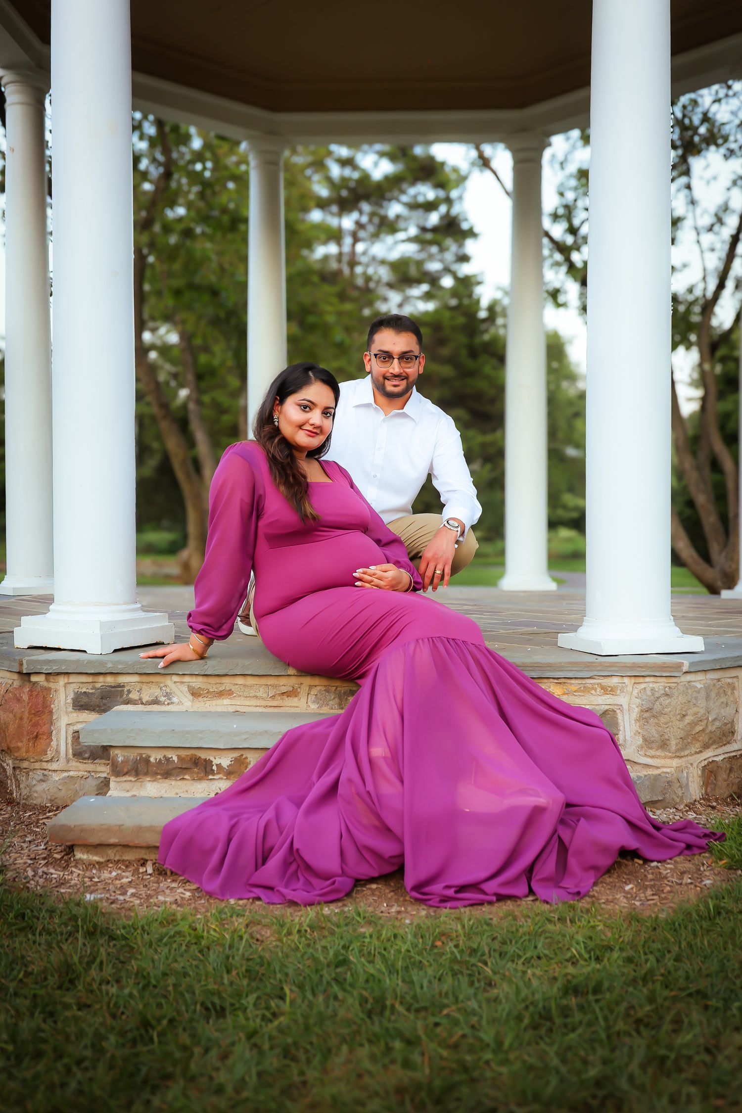 A couple poses together on a stone porch with white columns, the woman wearing a flowing magenta maternity gown.