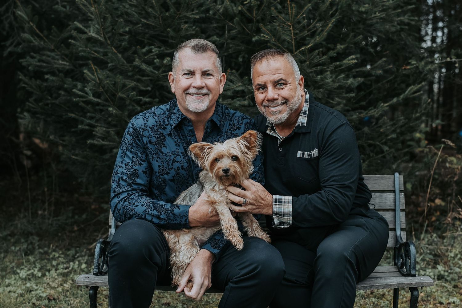 Two people sitting on a wooden bench outdoors with a small brown Yorkshire Terrier dog in their laps.