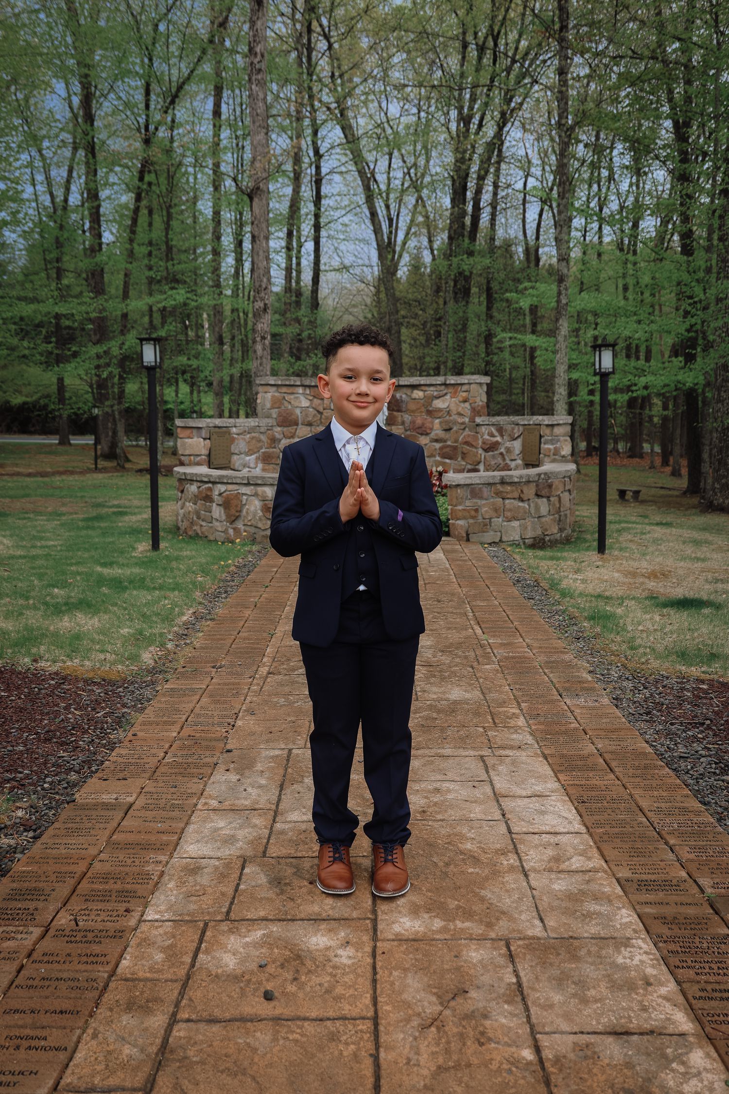 A young child in a navy suit walks and poses along a brick pathway surrounded by green trees and stone walls.