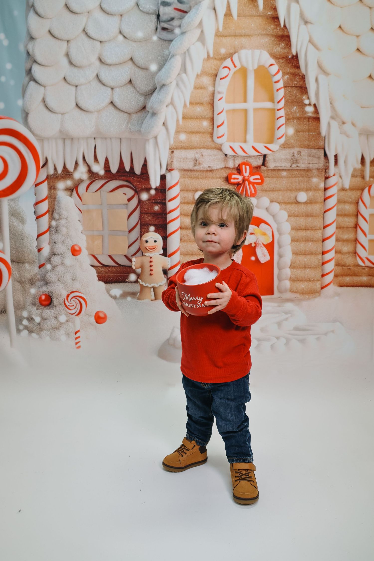 A small child in a red sweater and jeans stands in front of a whimsical candy-themed holiday backdrop with white decorations.