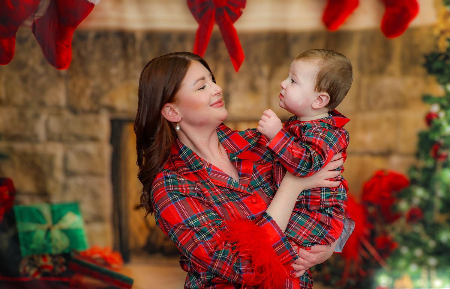 A pair in matching red plaid outfits pose together in front of a holiday backdrop with red bows and wrapped gifts.