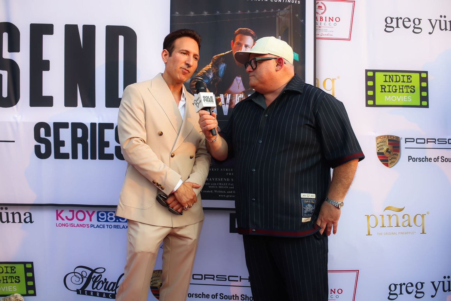 Two people in formal attire pose together at a media event with promotional banners and logos visible in the background.