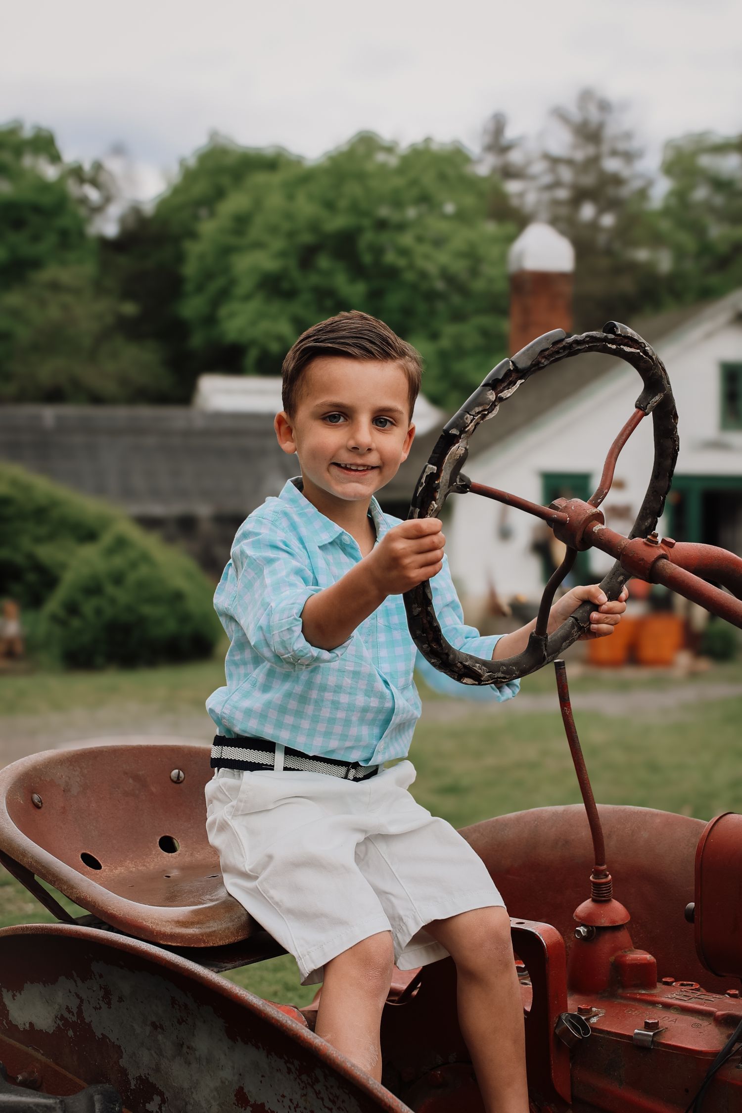 A young child in a plaid shirt and white shorts sits playfully on a vintage red tractor in a backyard setting.