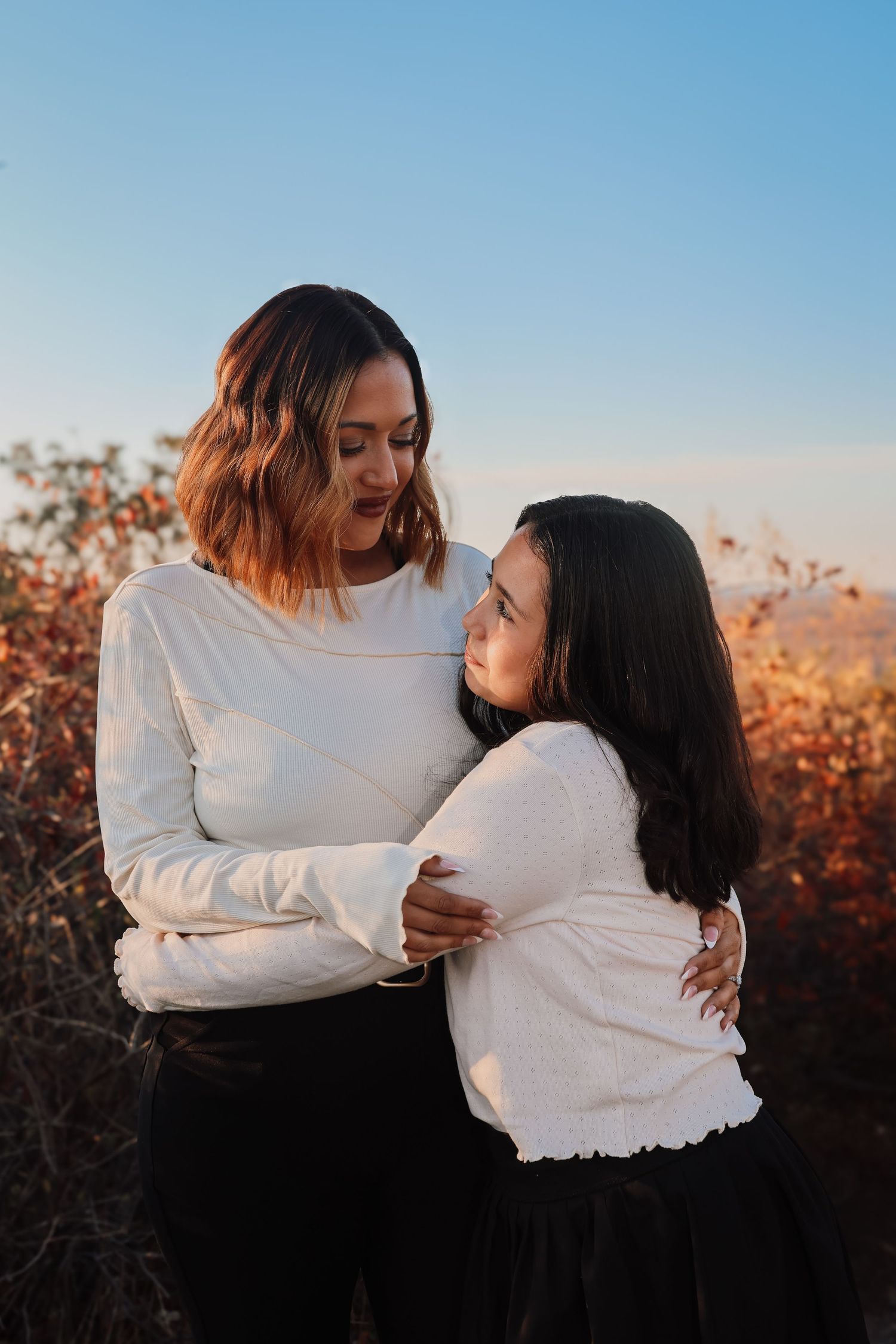 Two people in white sweaters embrace and share a tender moment against an autumn sunset backdrop.