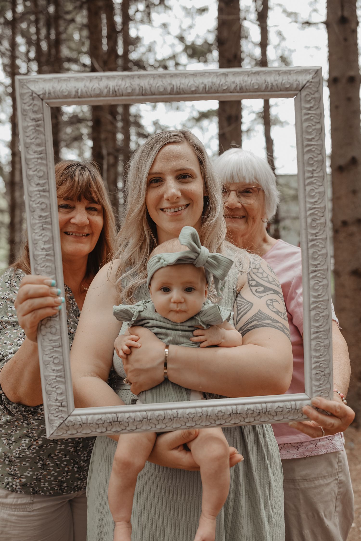 A multi-generational family portrait taken outdoors with a decorative white frame being held around them in a forest setting.