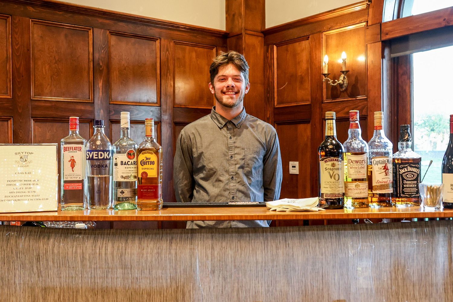 A bartender stands behind a wooden bar counter with various bottles of liquor and spirits displayed on the shelf.