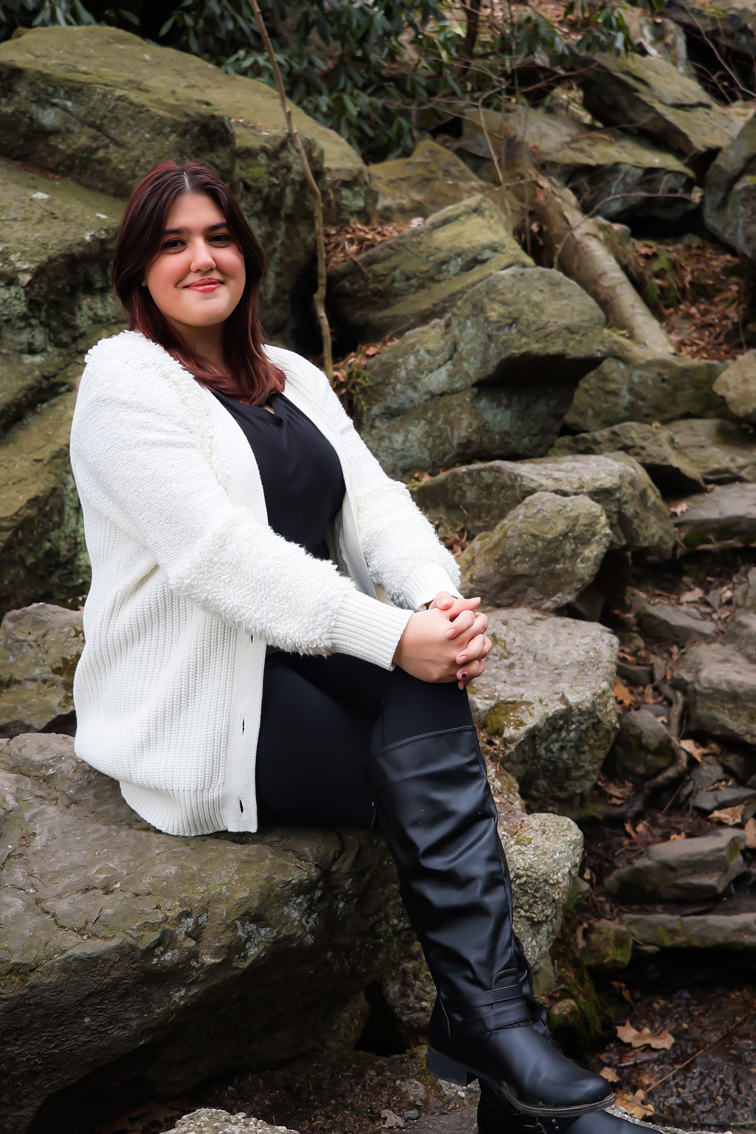 A person in a white sweater and black pants sits casually on rocky terrain outdoors.