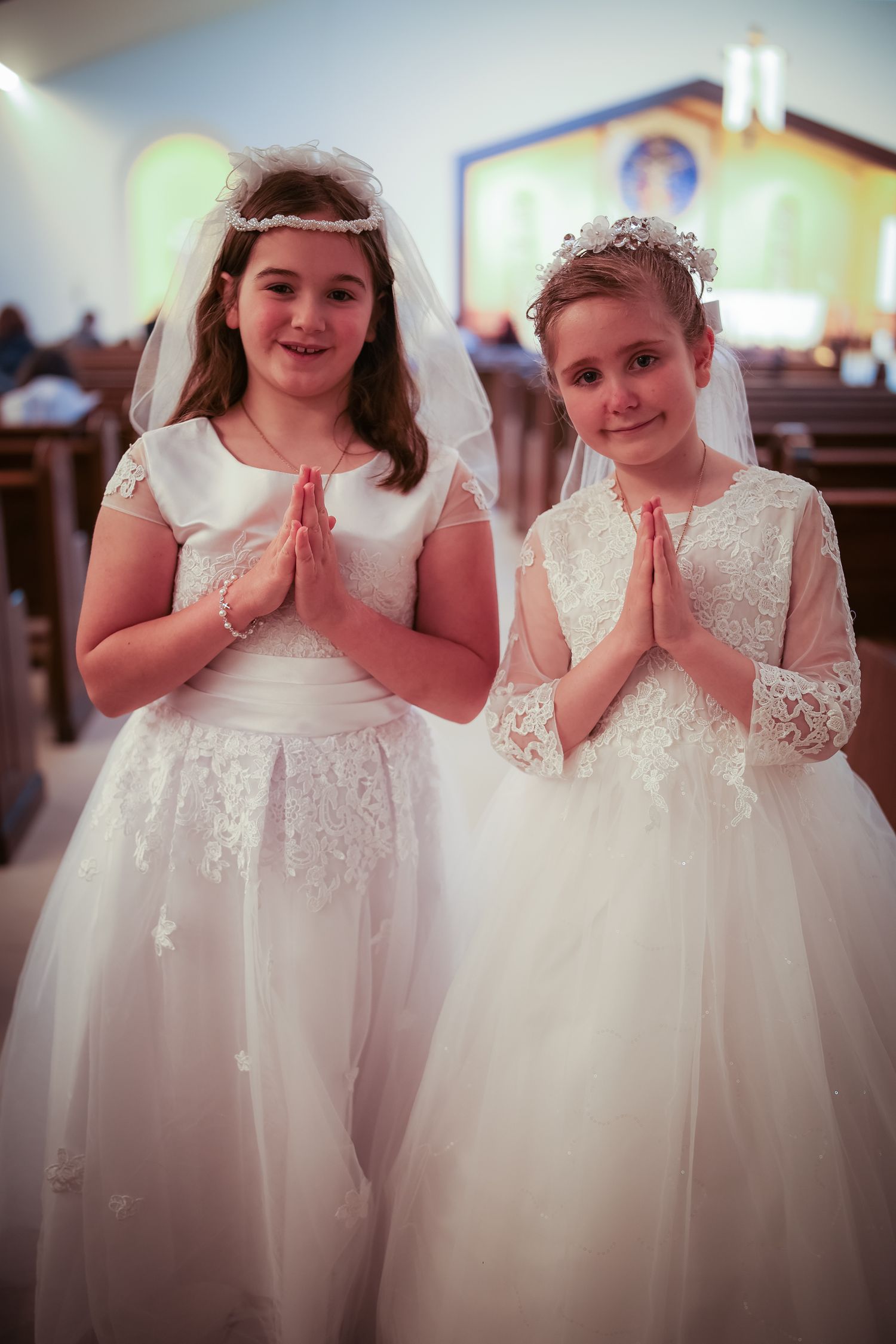 Two young flower girls in white lace dresses stand together clasping their hands in prayer at a formal event.