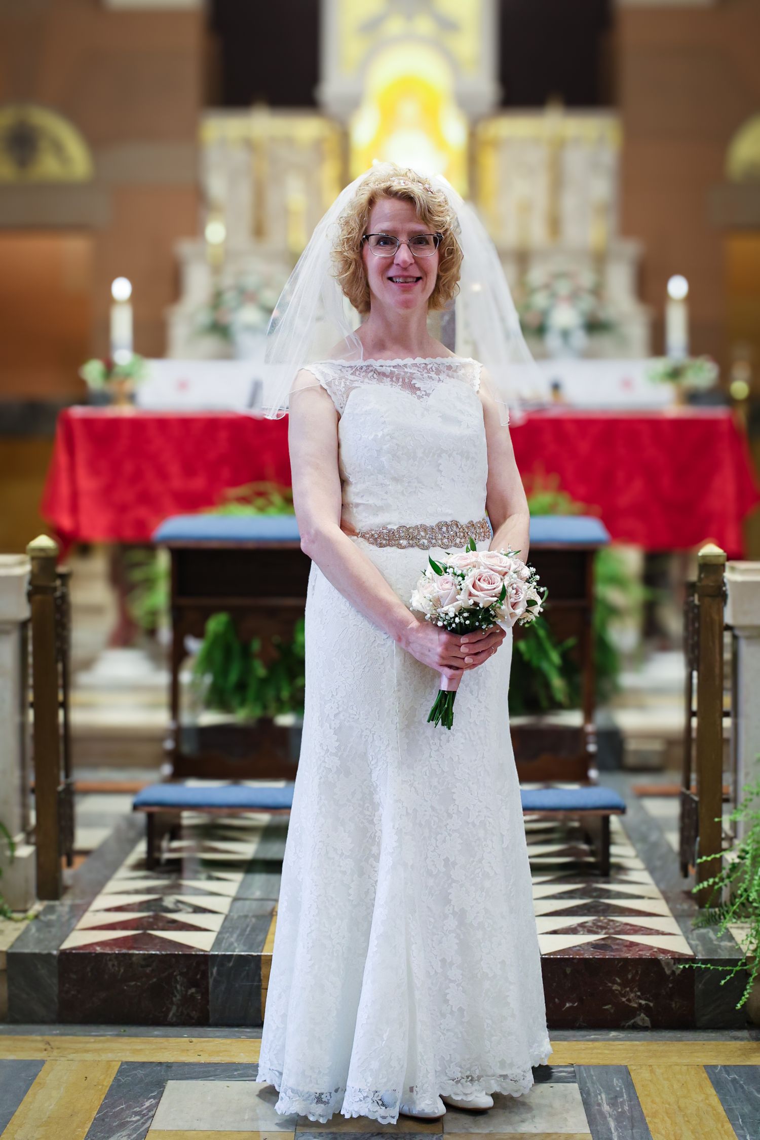 A bride in a white lace wedding dress poses in a church aisle holding a pink and white floral bouquet.