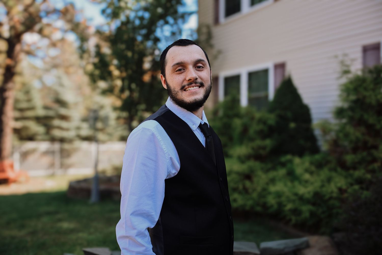 A groom in a black vest and white shirt smiles while standing outside a house during a wedding photoshoot.
