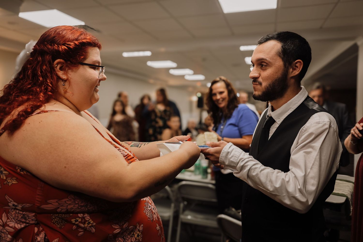 Two people exchange items during an indoor gathering while others watch in the background.