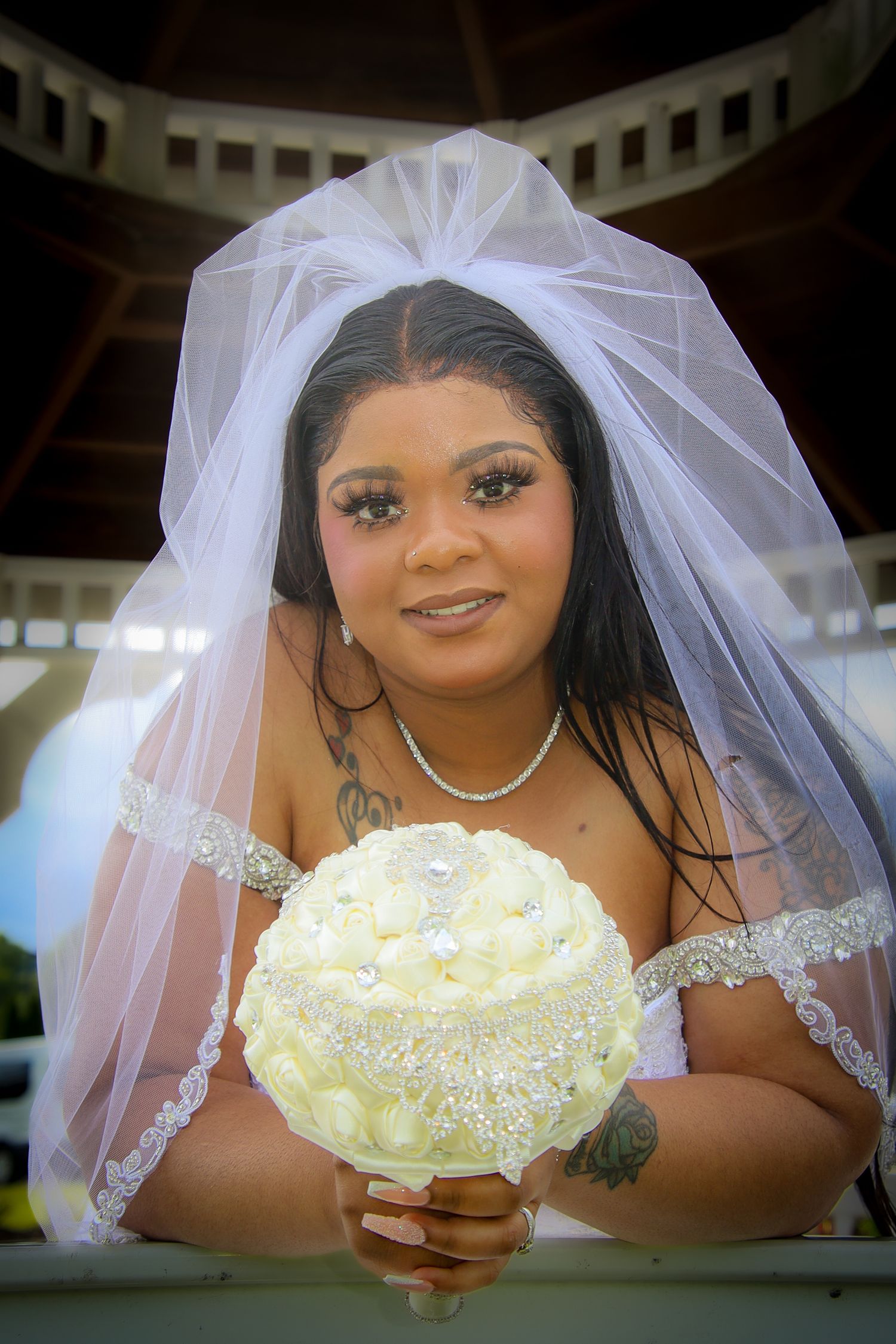 A bride in a white veil holds a cream-colored bouquet while posing outdoors at a gazebo.