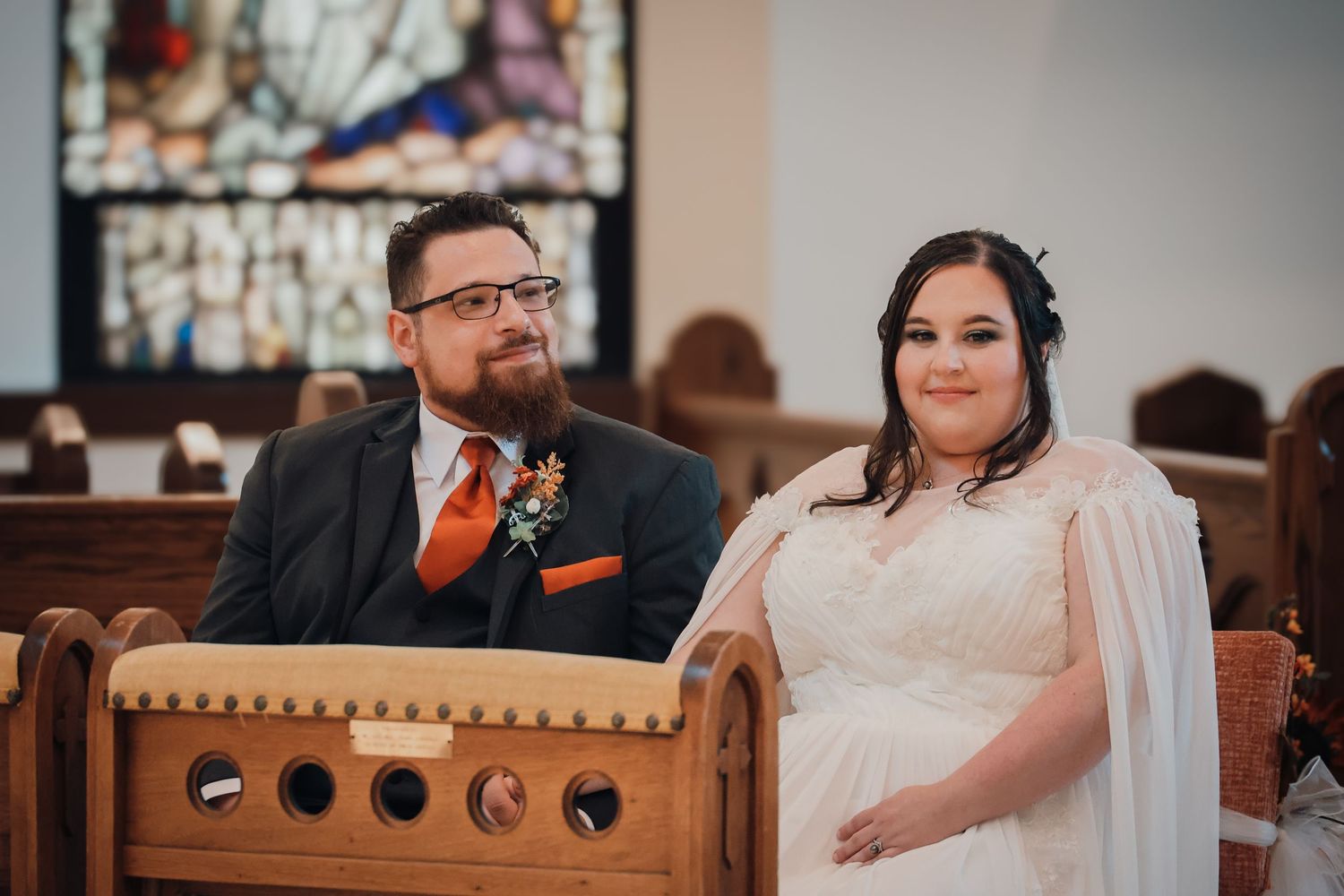 Newlywed couple sits together in ornate wooden church pew during wedding ceremony.
