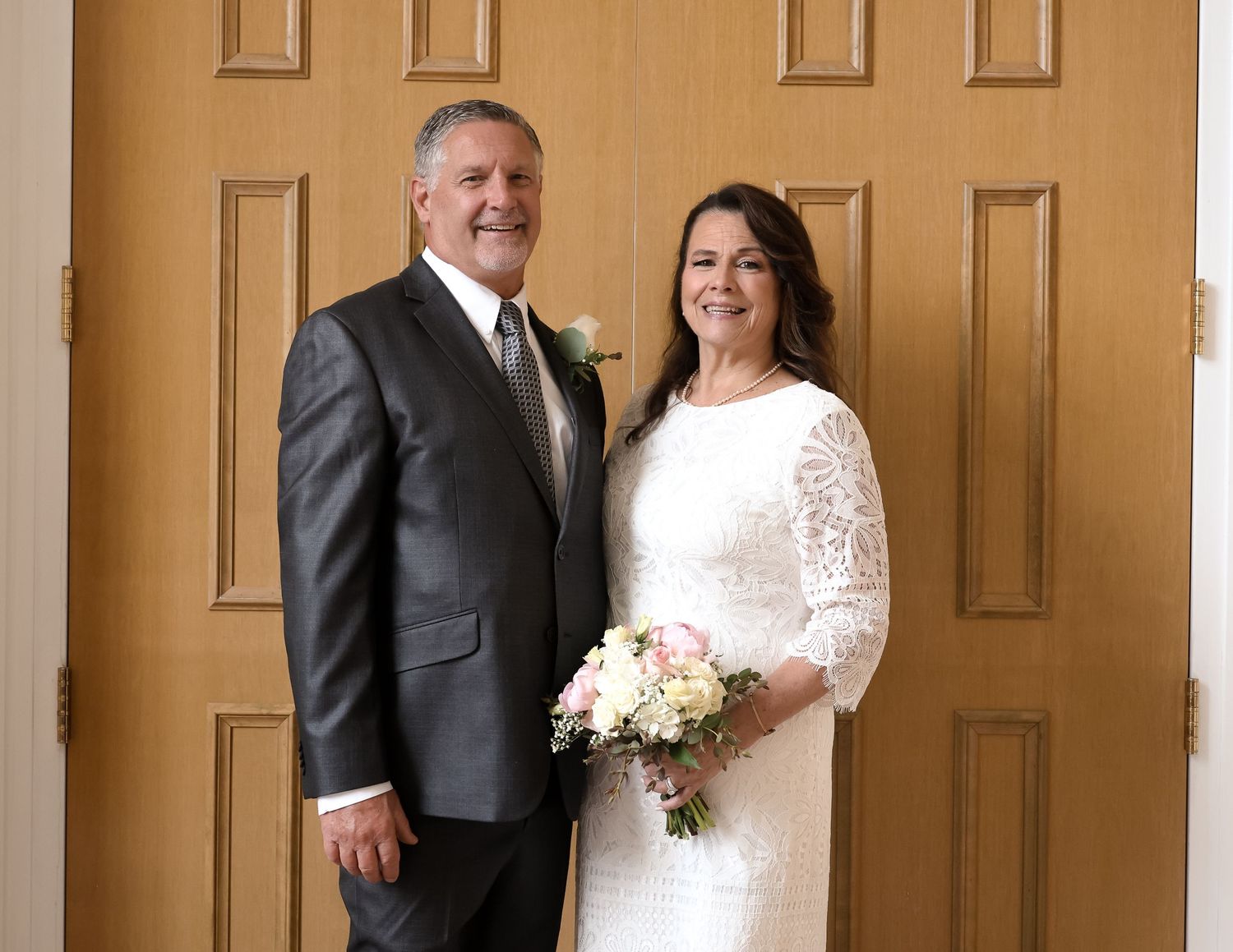 A couple poses together in formal wedding attire with a white bouquet against wooden doors.