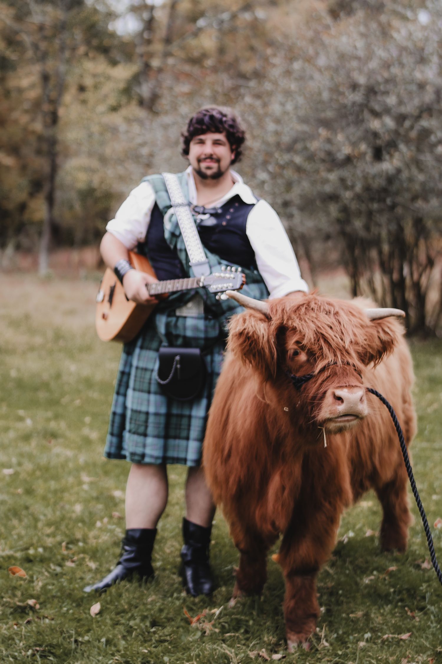 A person wearing traditional Scottish attire poses with a Highland cow in a pastoral field.