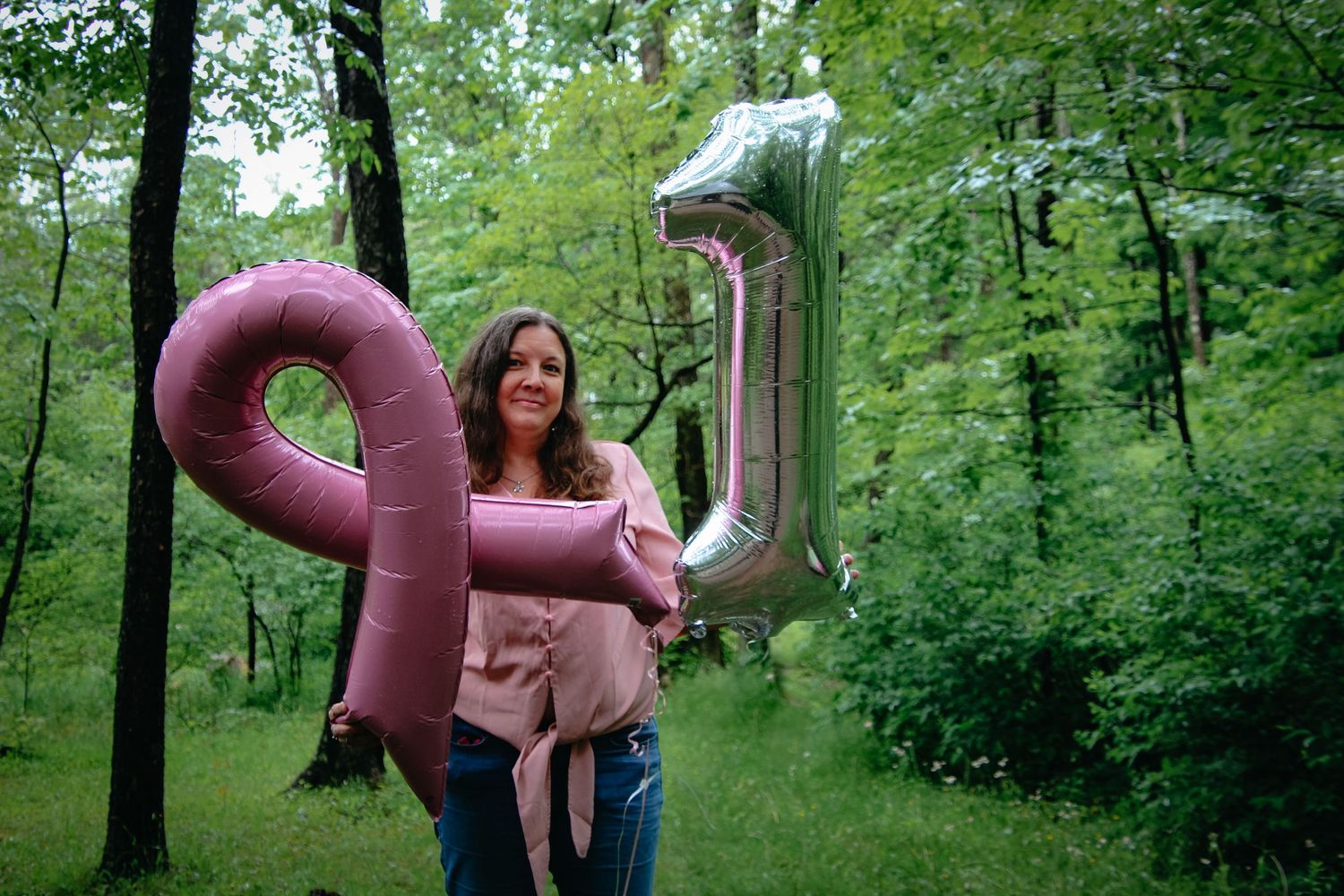 Someone holds pink and silver number one balloons in a lush green forest setting during a photo shoot.