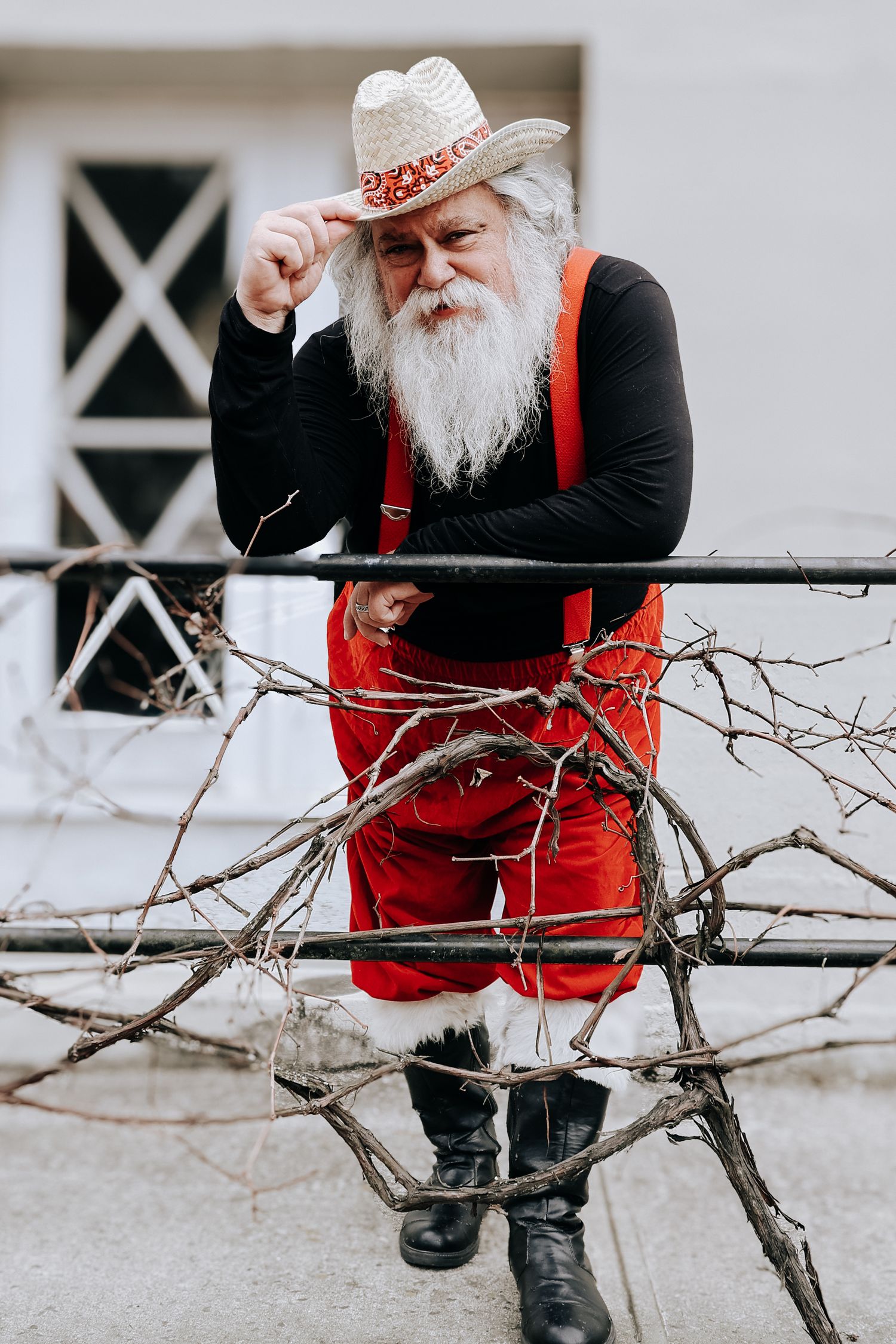 A person in red overalls and white beard sits on a porch railing while examining bare tree branches in winter.