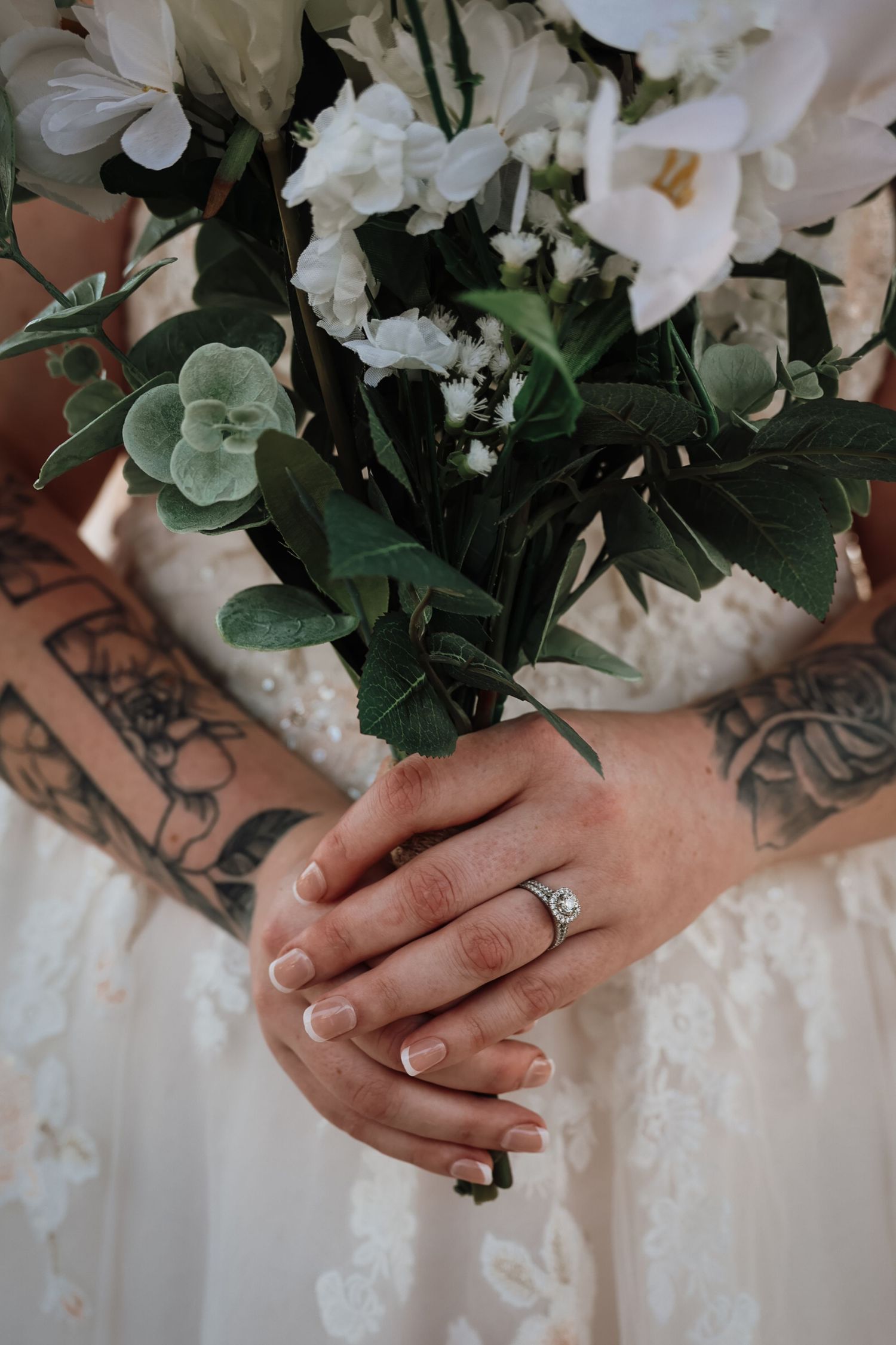Bride holding white floral bouquet with green leaves, wearing diamond engagement ring, with tattooed arms visible.