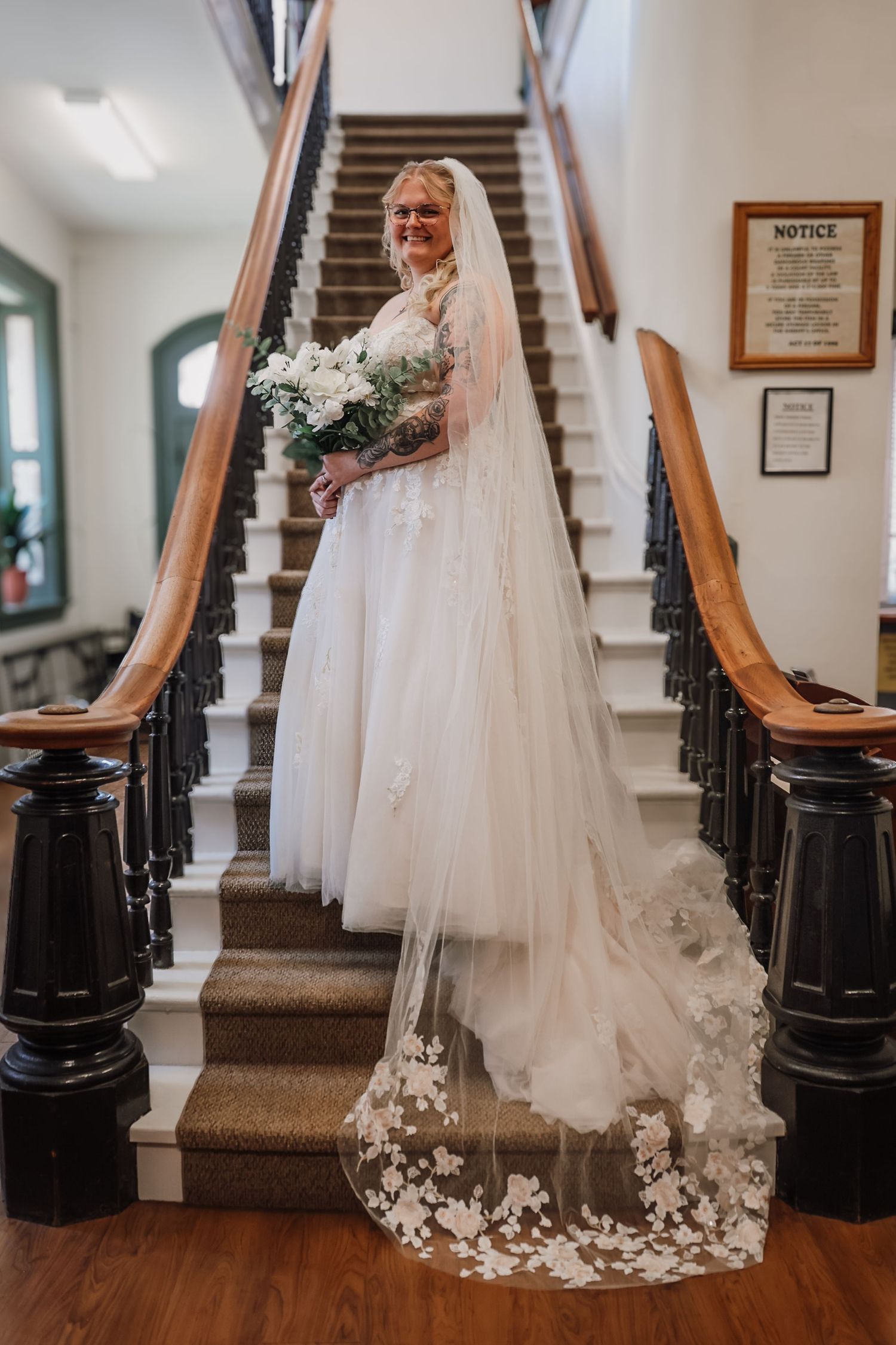 Bride in a white tulle gown with floral veil holding a bouquet, posing on a grand staircase.