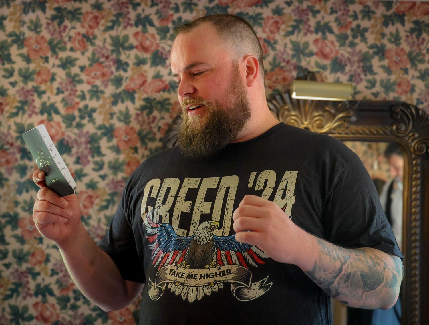 Bearded man in a Creed 24 black t-shirt smiling while looking at a card in a floral wallpaper room.