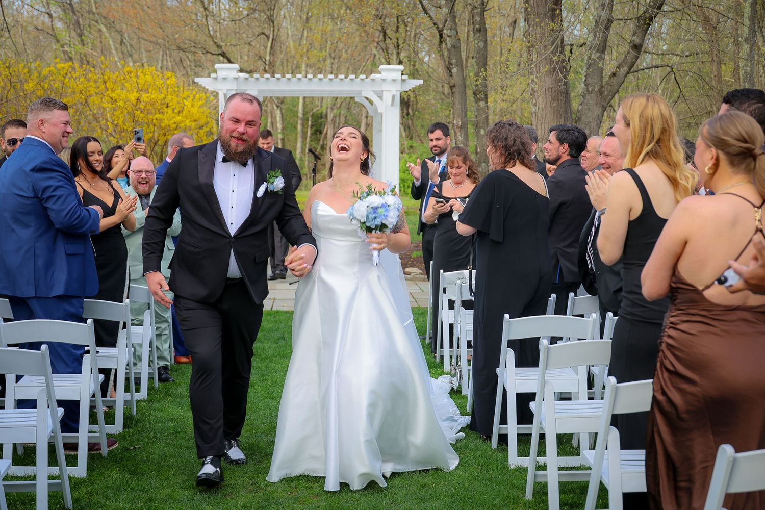 Joyful newlyweds walk down the aisle after outdoor wedding ceremony, guests applauding, white chairs, floral arbor backdrop.