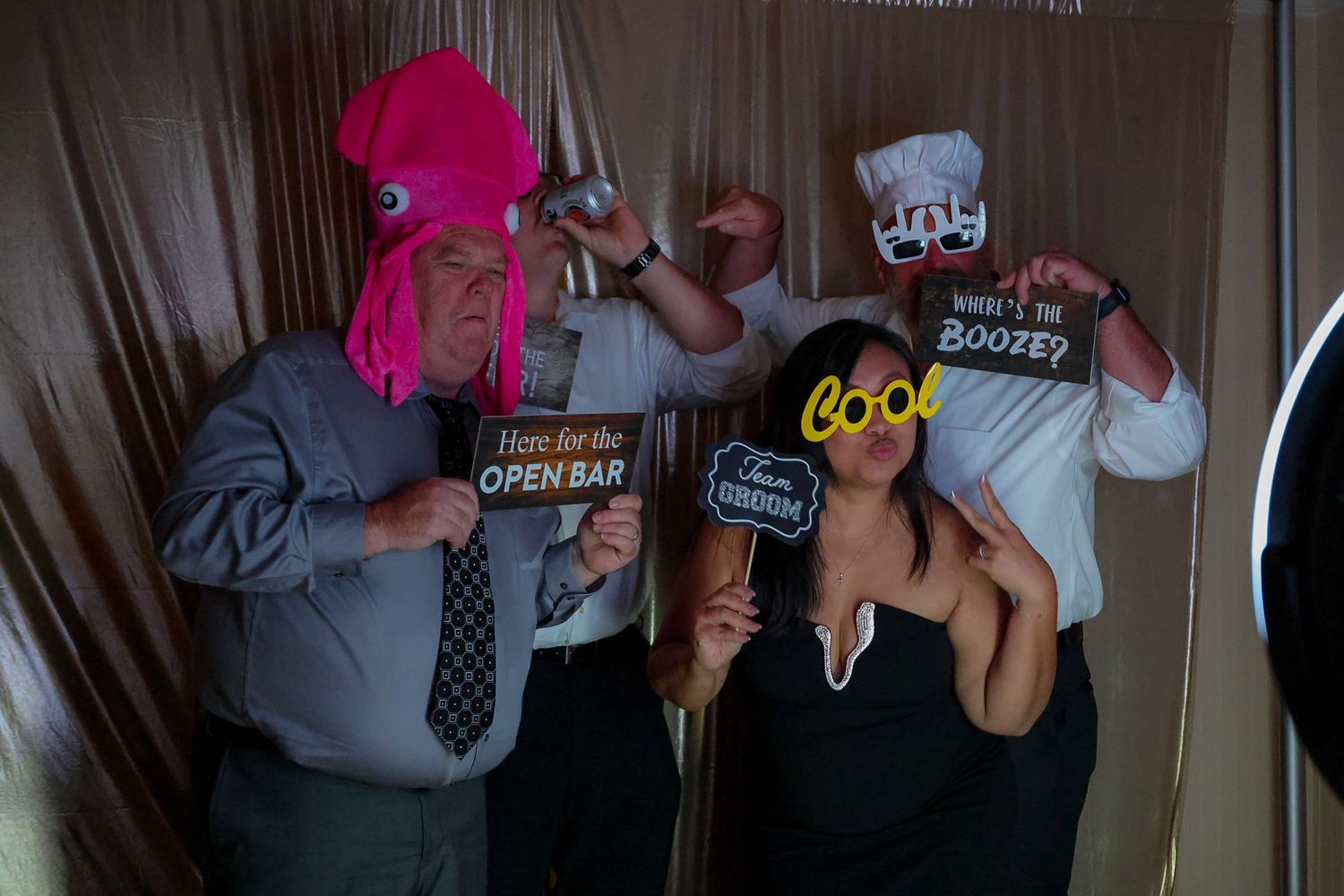 People posing with fun photo booth props including a pink octopus hat and signs reading Here for the Open Bar and Boozey.