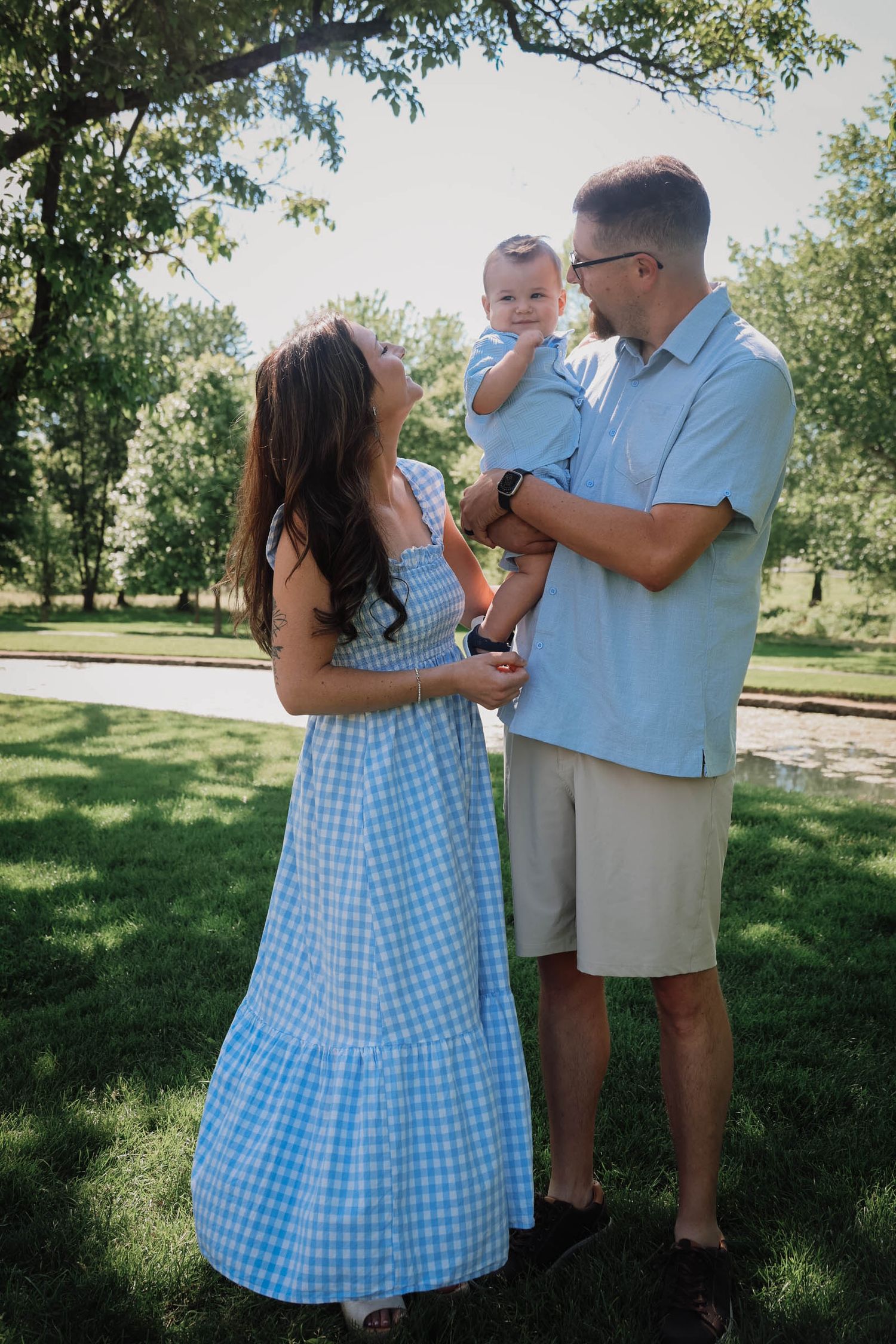Young family with baby boy in blue outfits pose together in sunny green park.