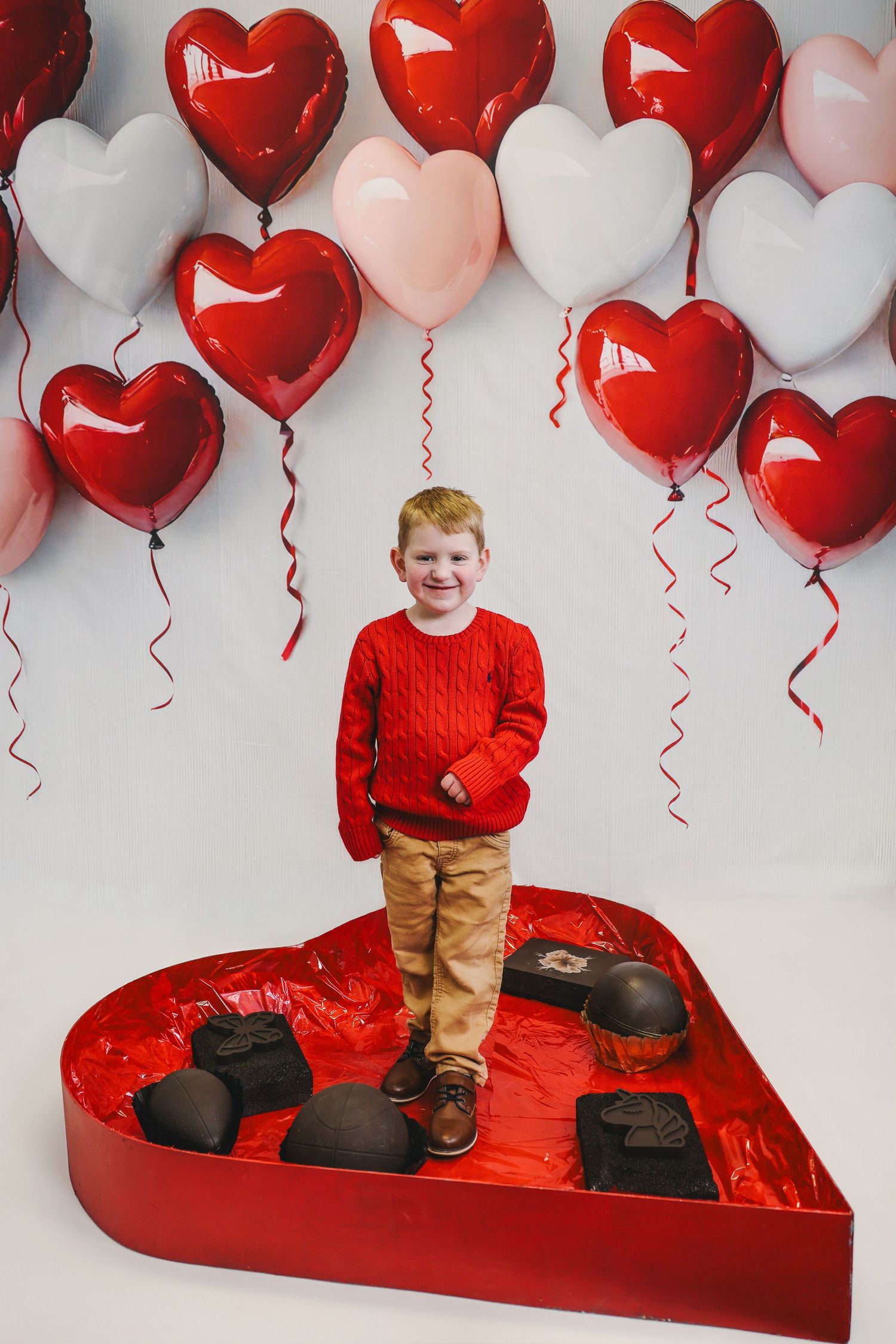 Smiling boy in red sweater standing in giant heart-shaped chocolate box with heart balloons backdrop.