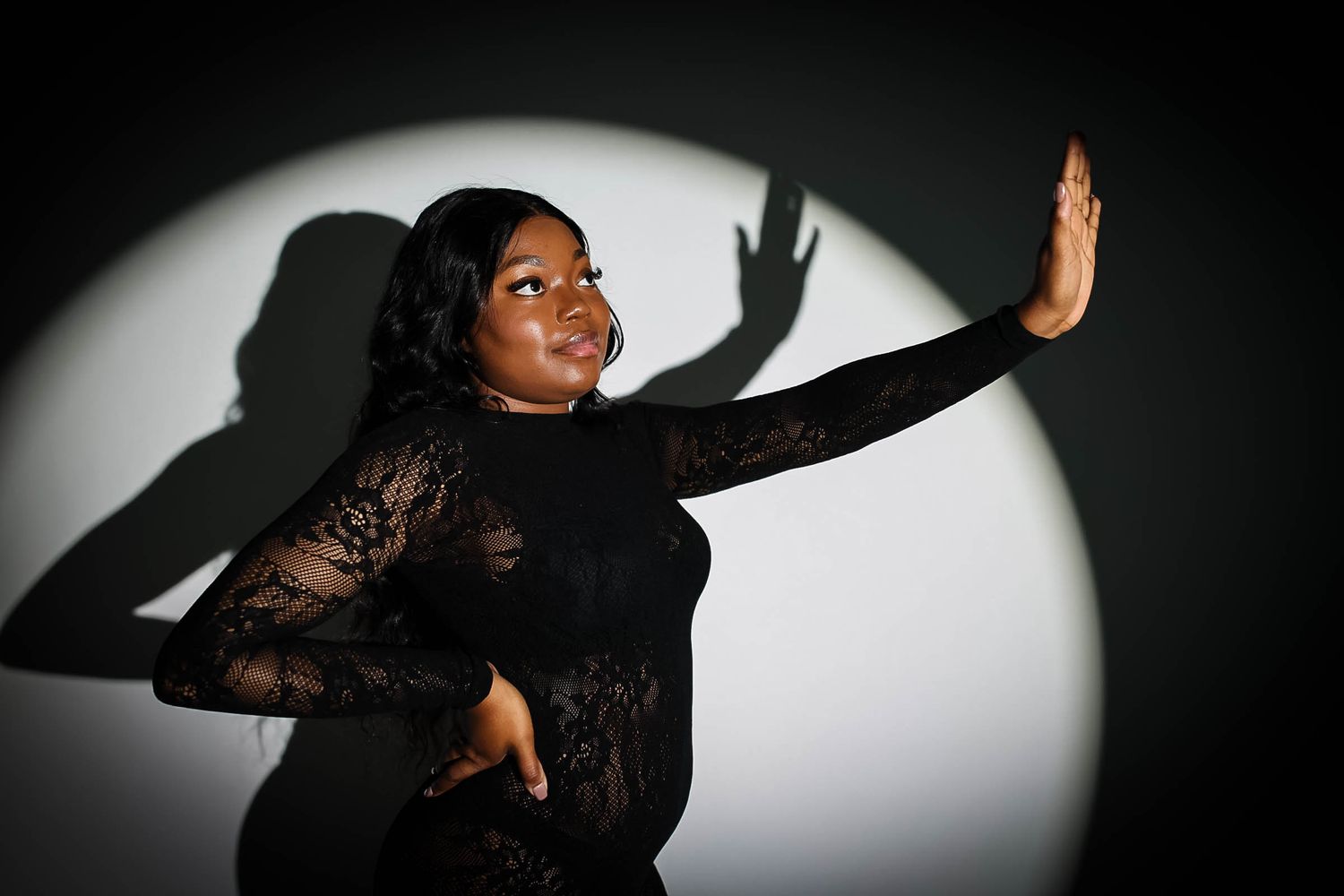 Woman in black lace dress poses dramatically against spotlight with expressive shadow on gray wall.